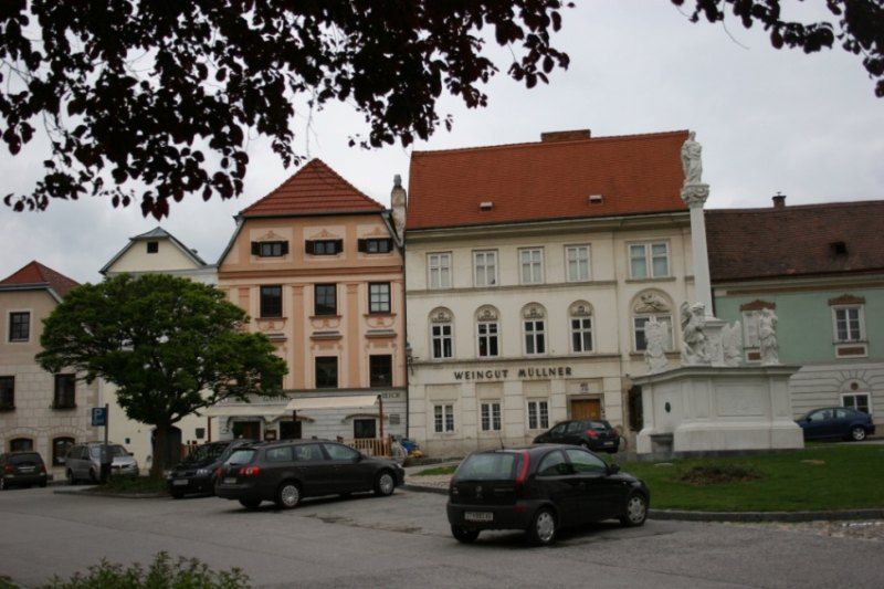Historic buildings with the sign 'Weingut Müllner' and a statue on a square.