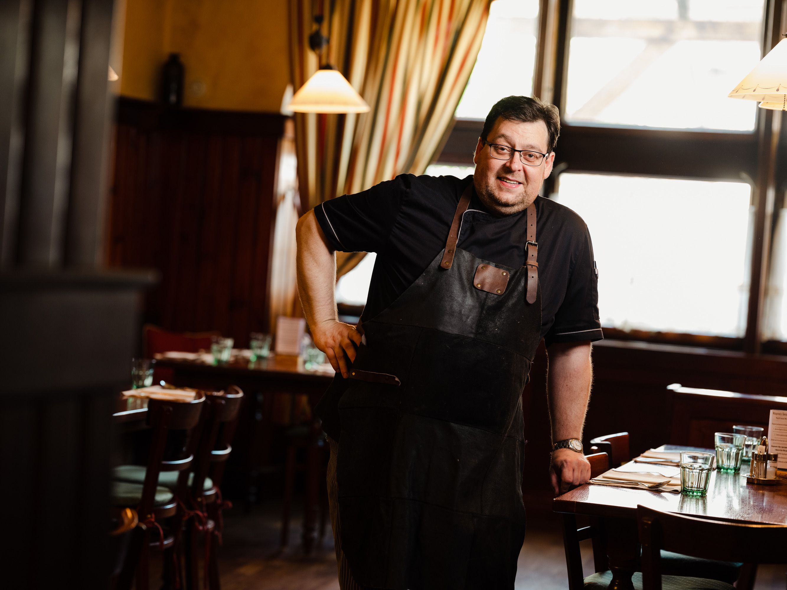 Man in a black apron stands smiling in a cozy restaurant.