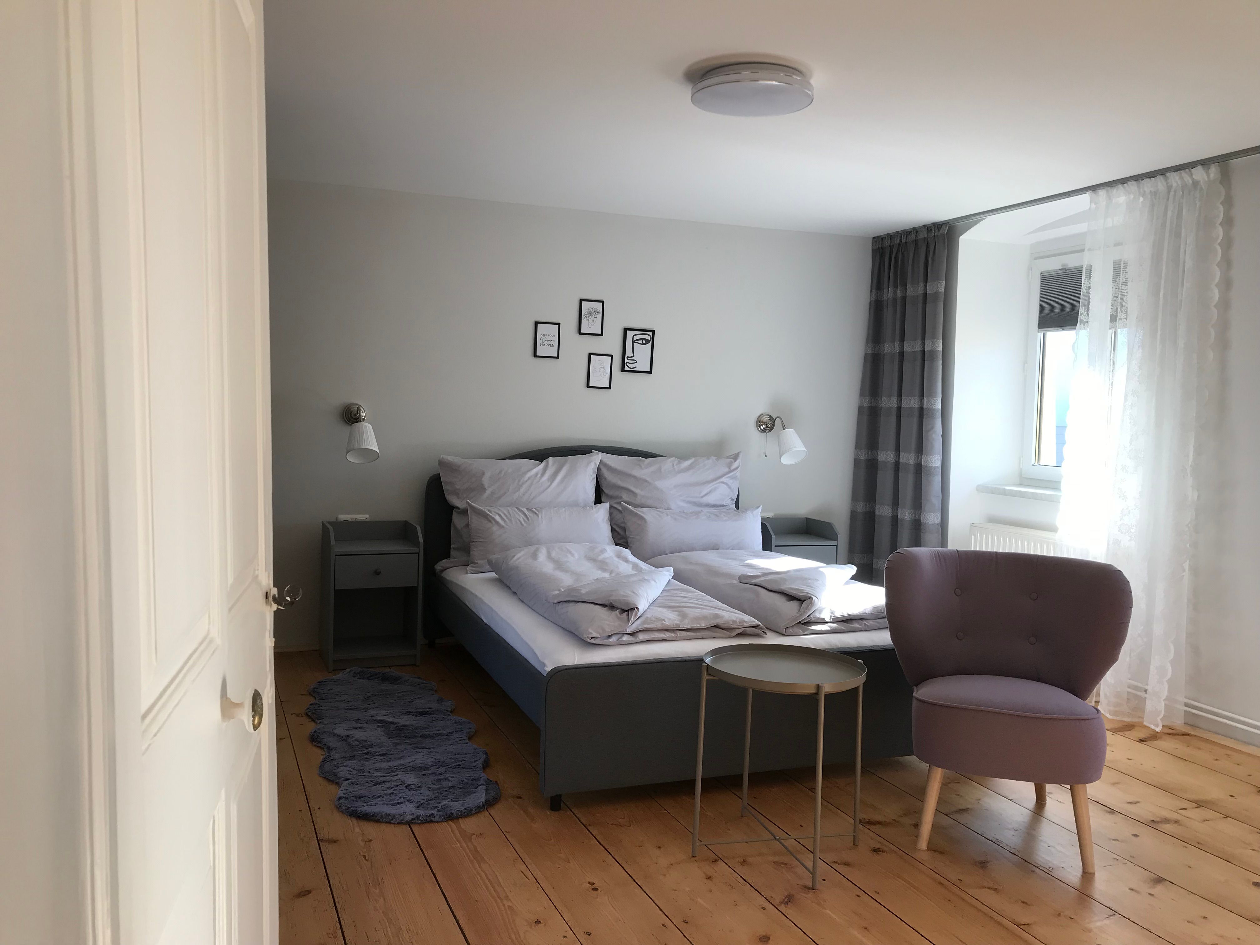 Modern furnished double room with bed, armchair and small table on wooden floor.