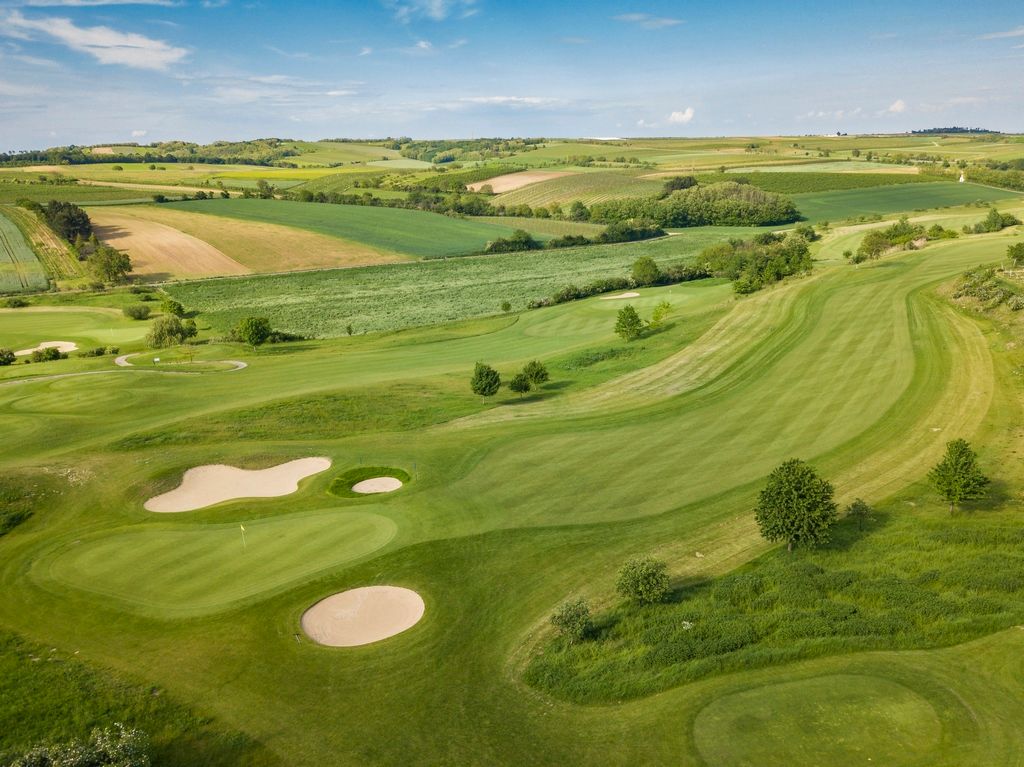 Aerial view of a golf course with green fairways and sand bunkers, surrounded by fields and hills.