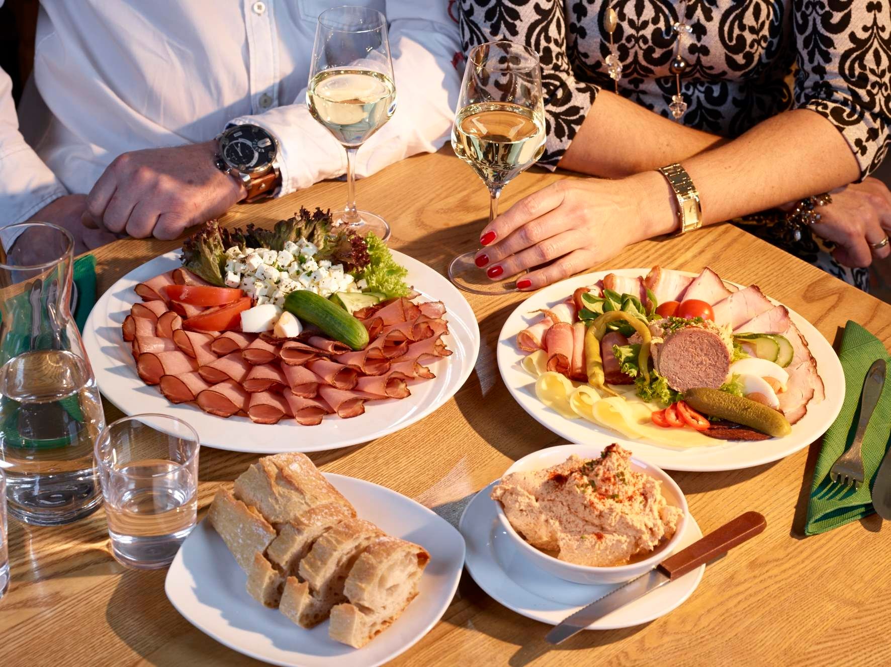 Two people sit at a table with platters of cold meats and cheese, bread and wine.