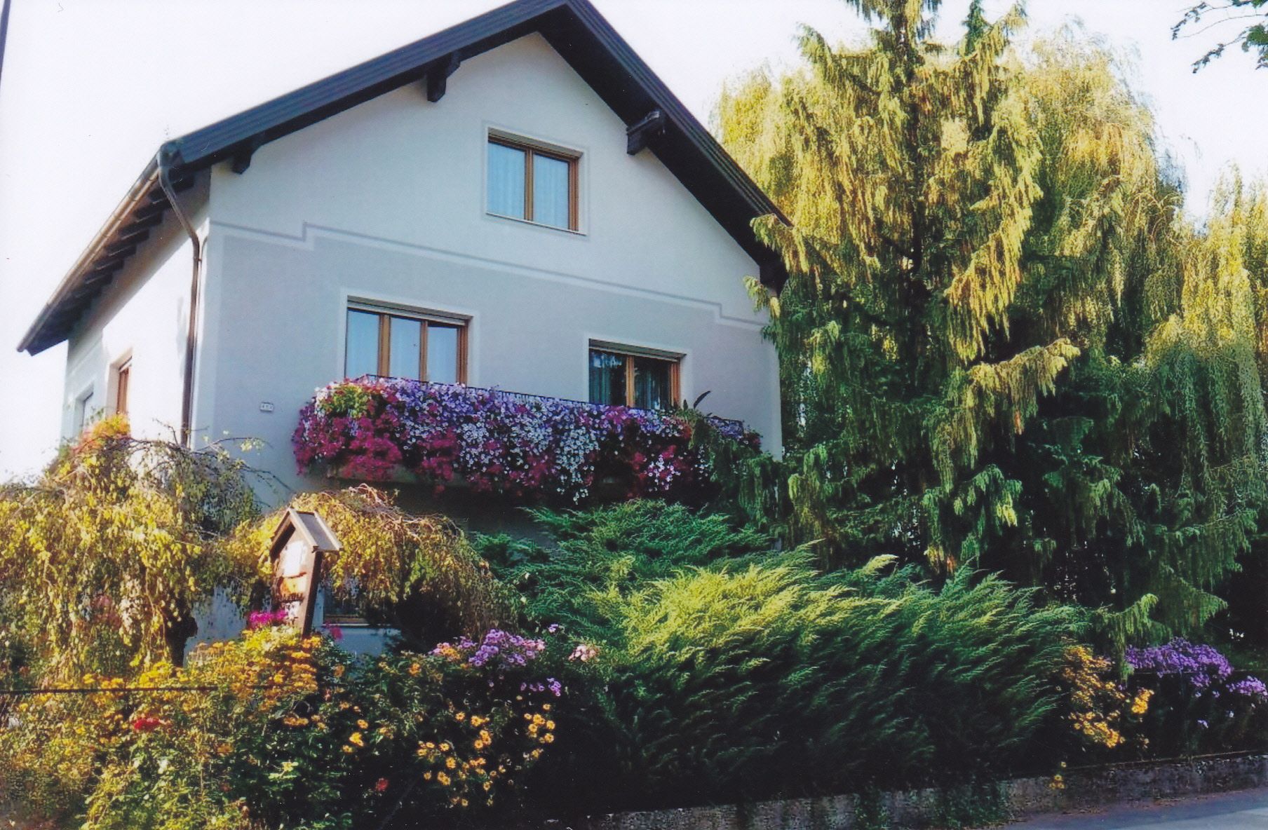 A two-story house with flowers on the balcony and surrounded by trees and shrubs.