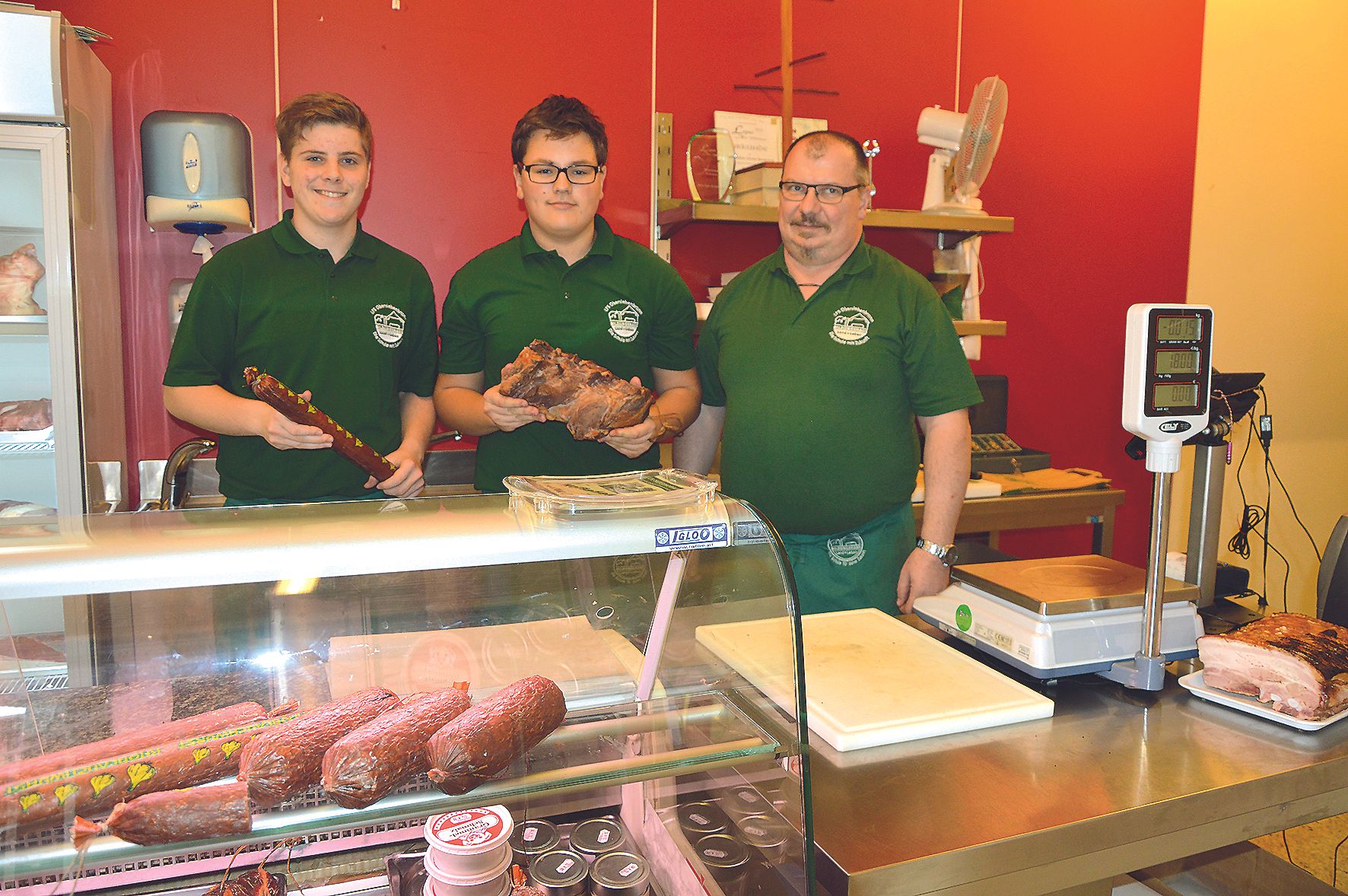 Three men in green shirts stand behind a meat counter in a farm store.