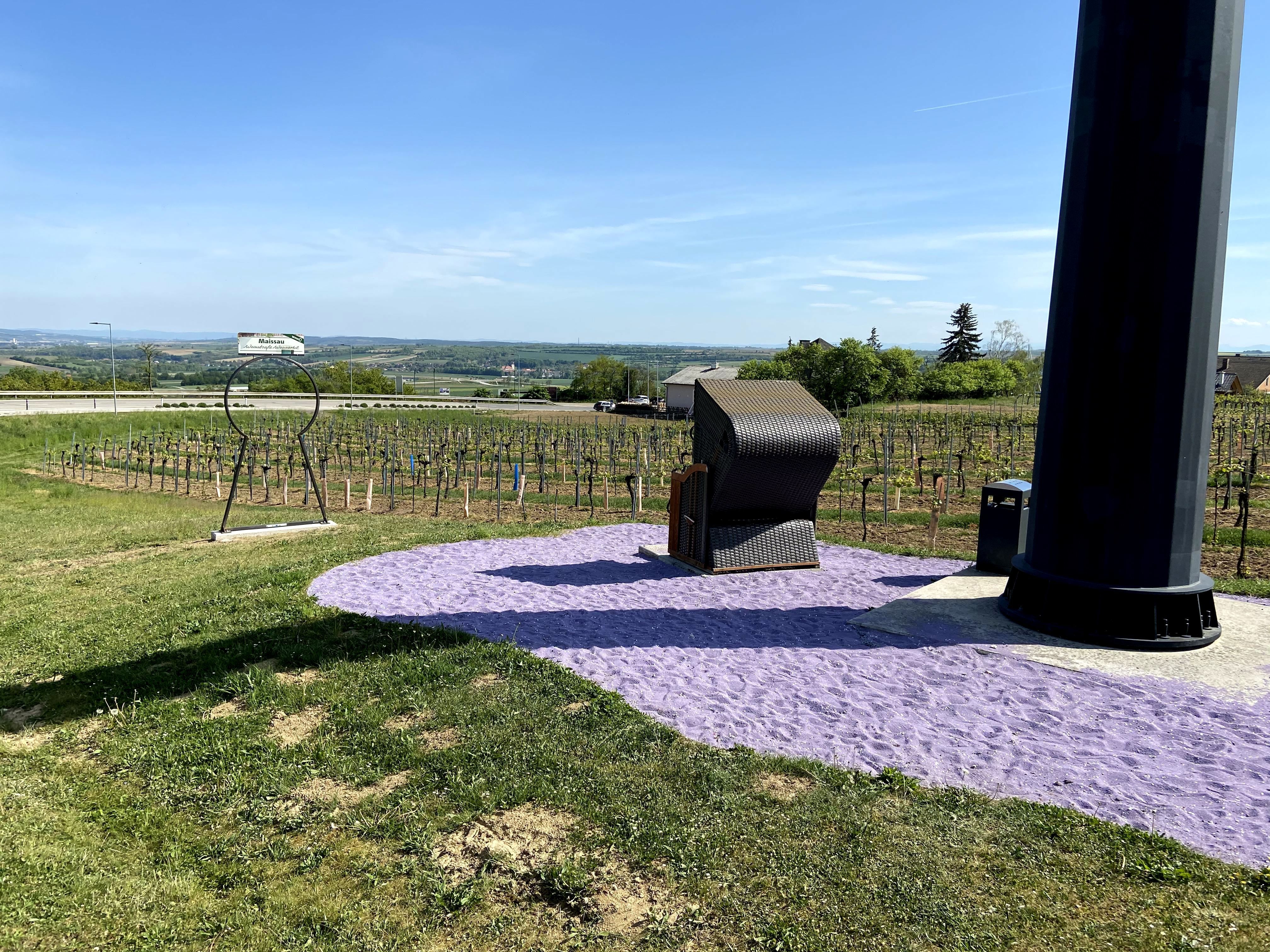 A beach chair stands on purple sand next to a large black pillar, surrounded by vineyards and a vast landscape.