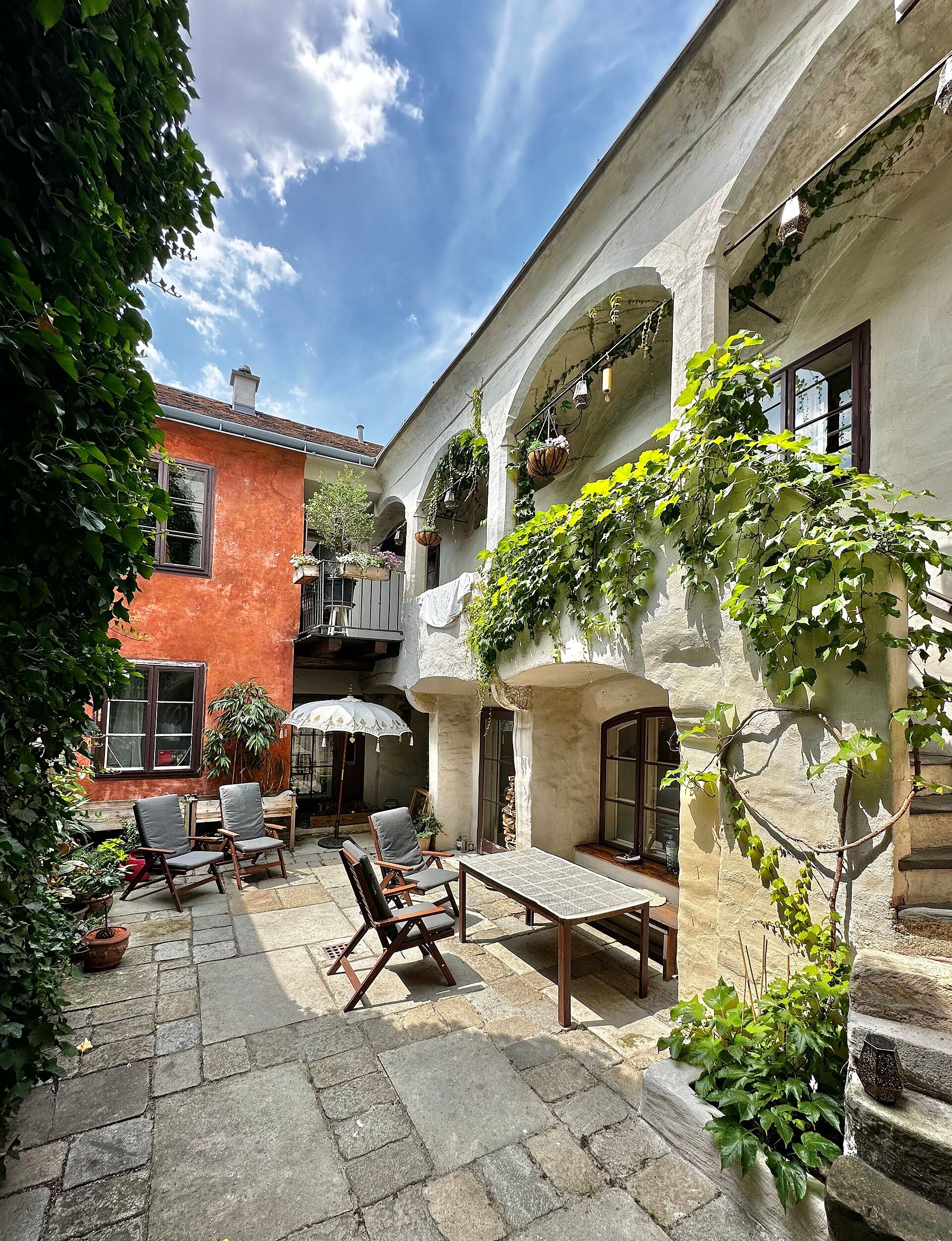 A cozy courtyard with plants, wooden furniture and a parasol.