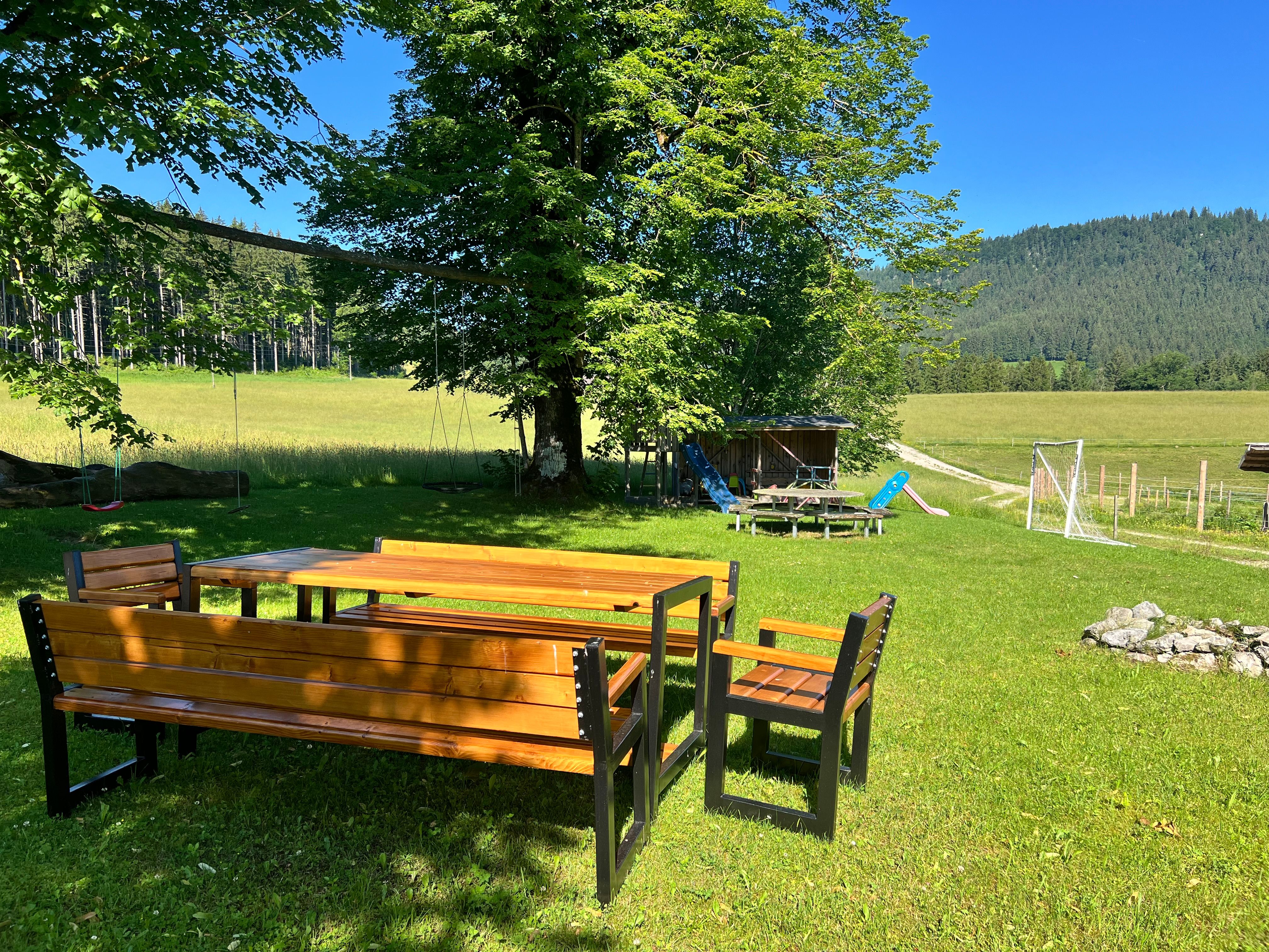 A garden with a wooden table and benches, surrounded by trees and meadows, with a playground in the background.