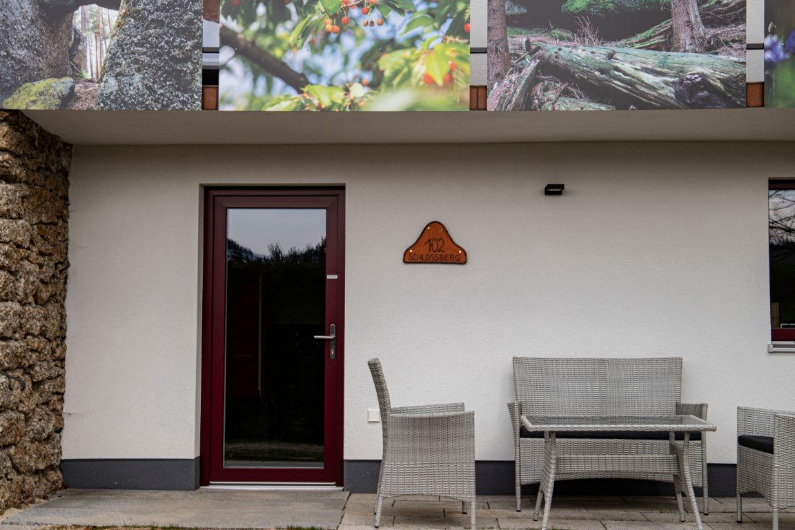 Entrance area of a building with a door, a sign with the inscription '102 Schlossberg' and garden furniture in front of it.