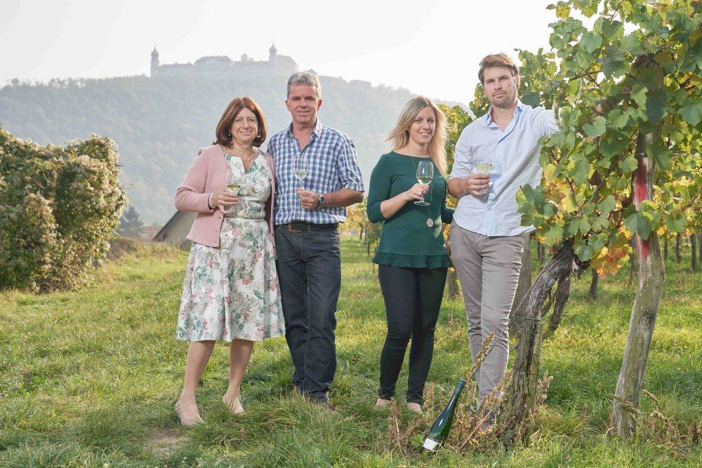 Four people are standing in a vineyard with wine glasses in their hands; a castle on a hill can be seen in the background.