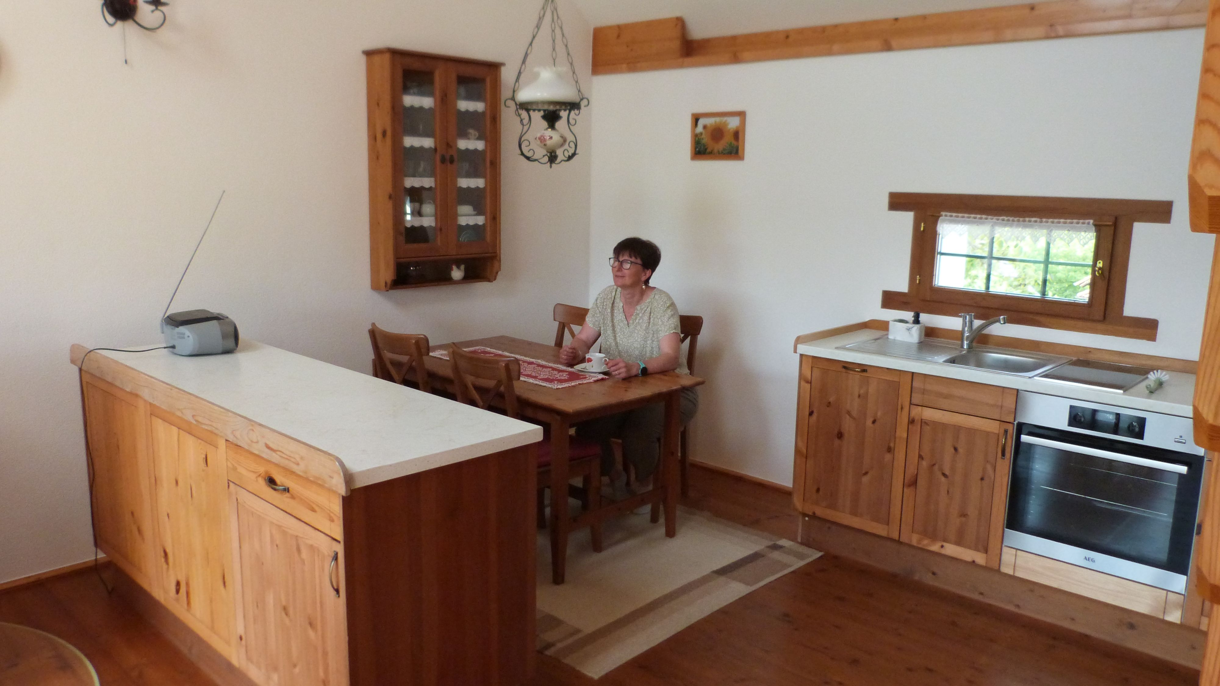 A woman sits at a wooden table in a cozy kitchen with wooden cupboards and a small window.