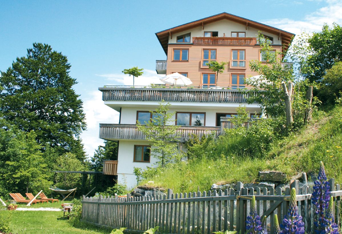 A multi-storey hotel with wooden balconies, surrounded by green nature and blooming flowers, under a blue sky.