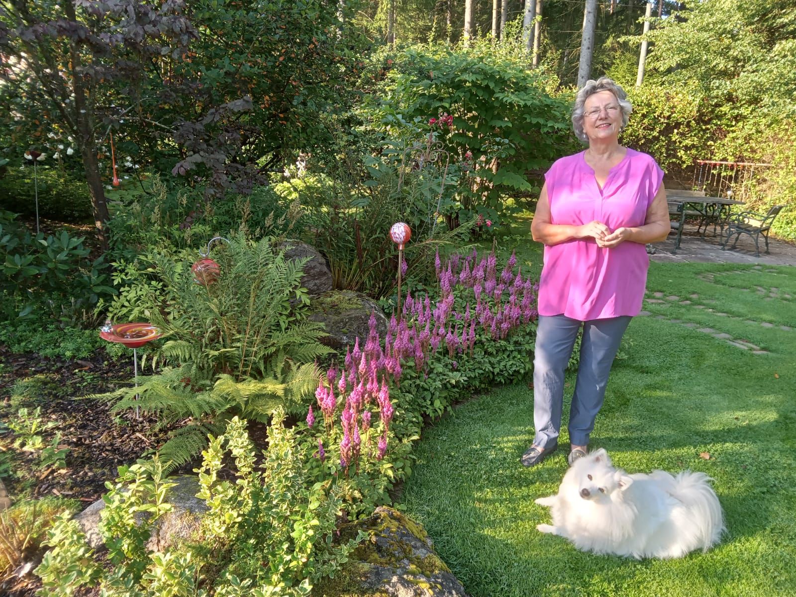 A woman stands in a garden next to a white dog on the lawn.