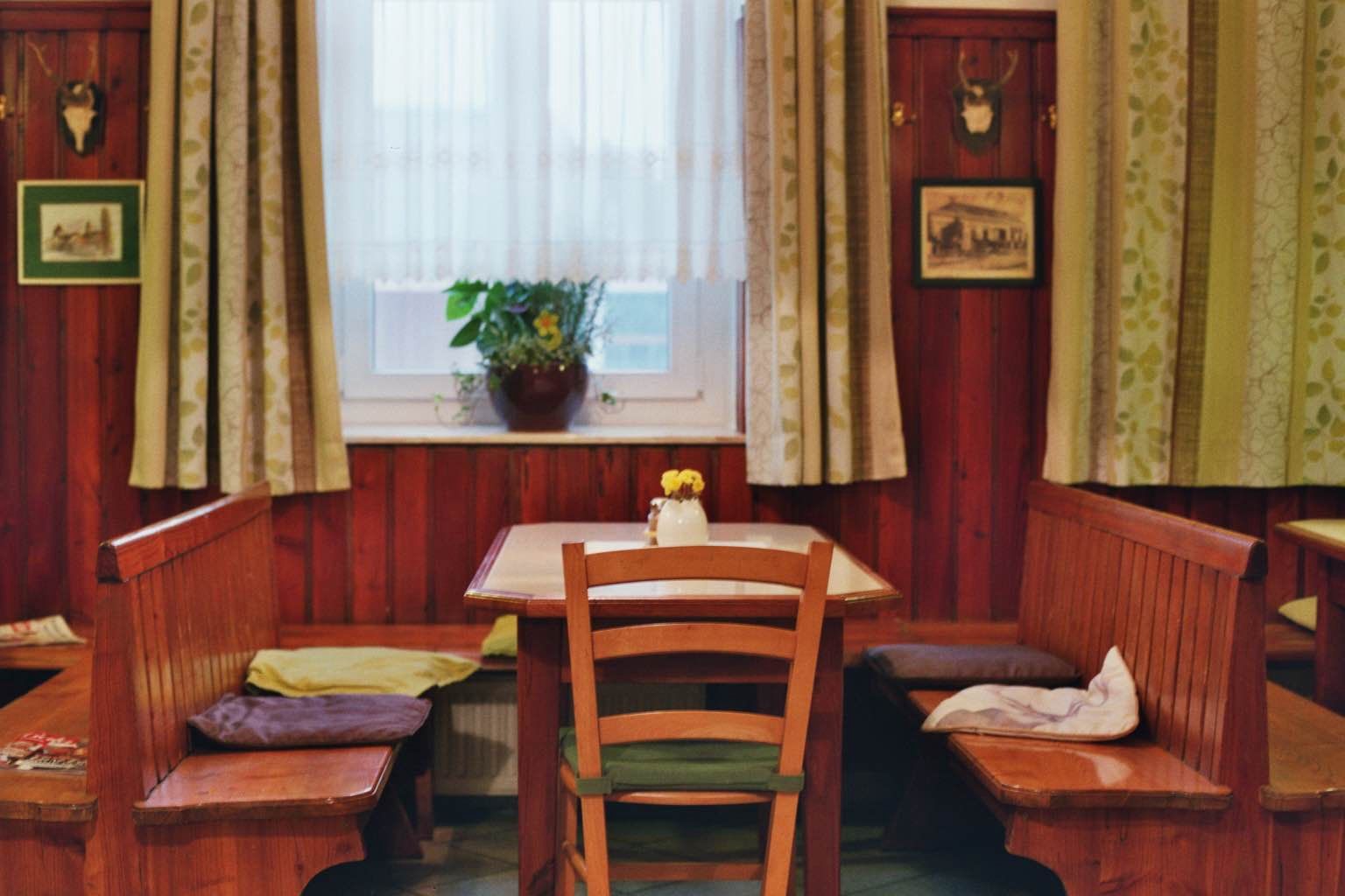 Cozy dining room with wooden furniture, table, chairs and bench, windows with curtains, plants and wall decorations.