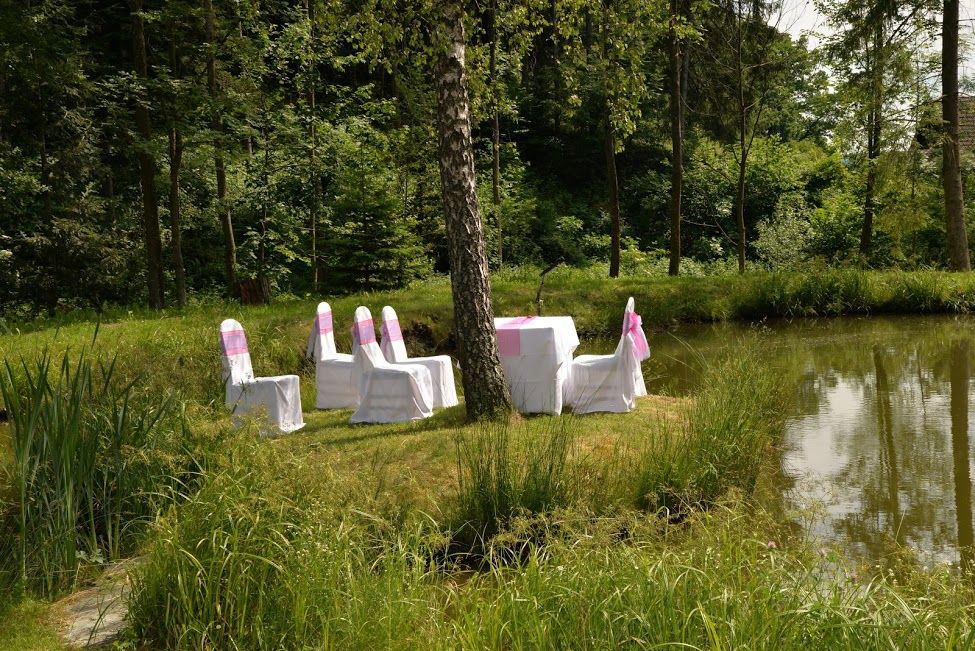 Chairs and a table with white covers on a green meadow next to a pond, surrounded by forest.