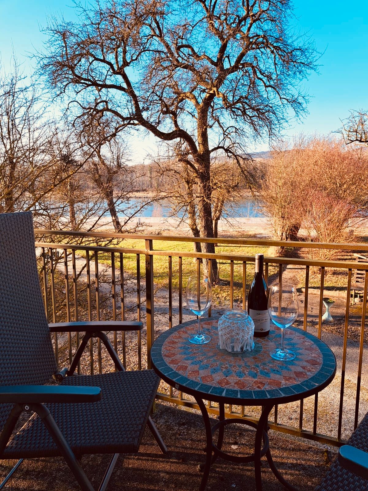 Balcony with table, two wine glasses and wine bottle, view of trees and river.
