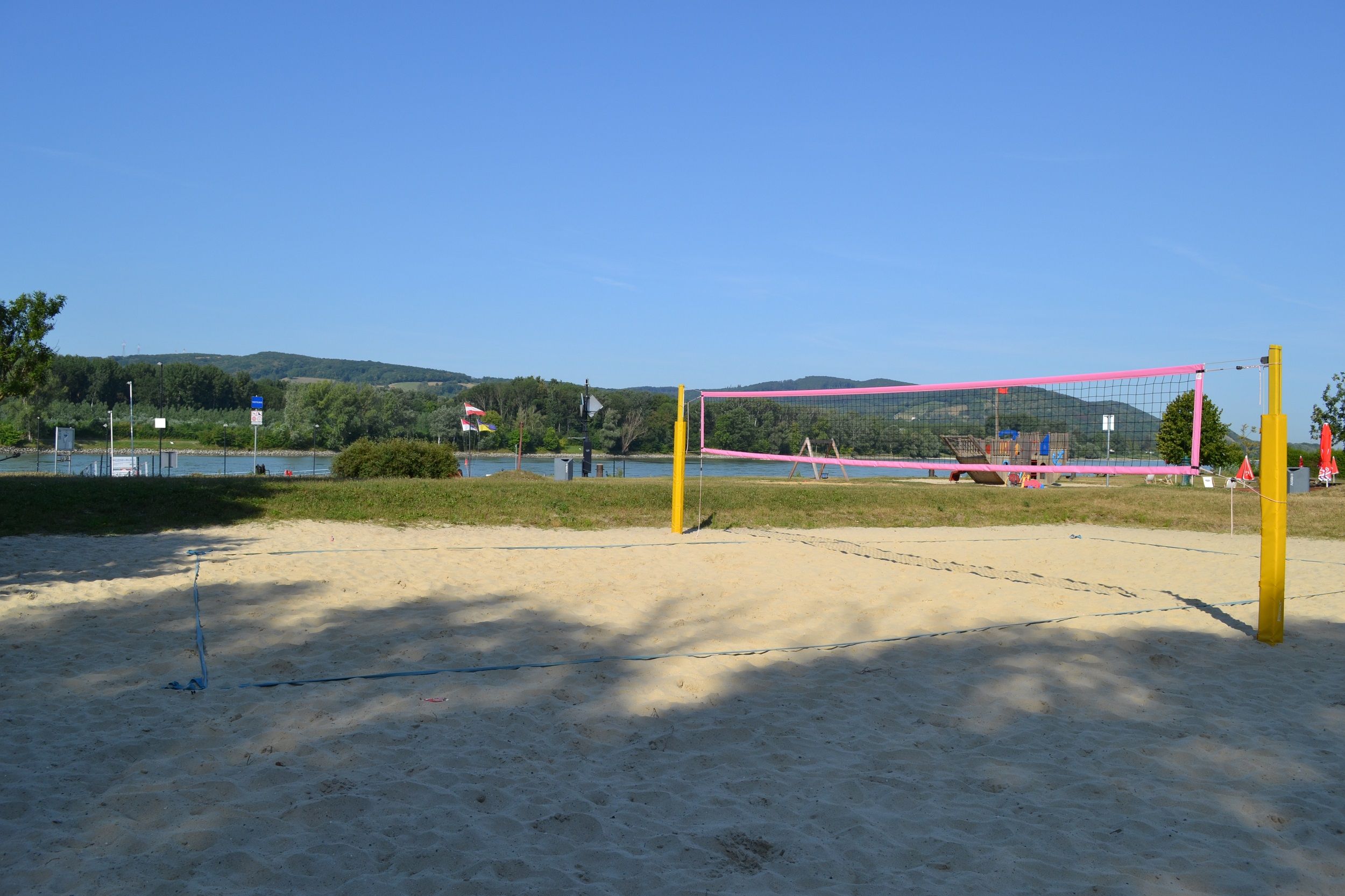 An empty beach volleyball court with yellow posts and a pink net in front of a river and green hills in the background.