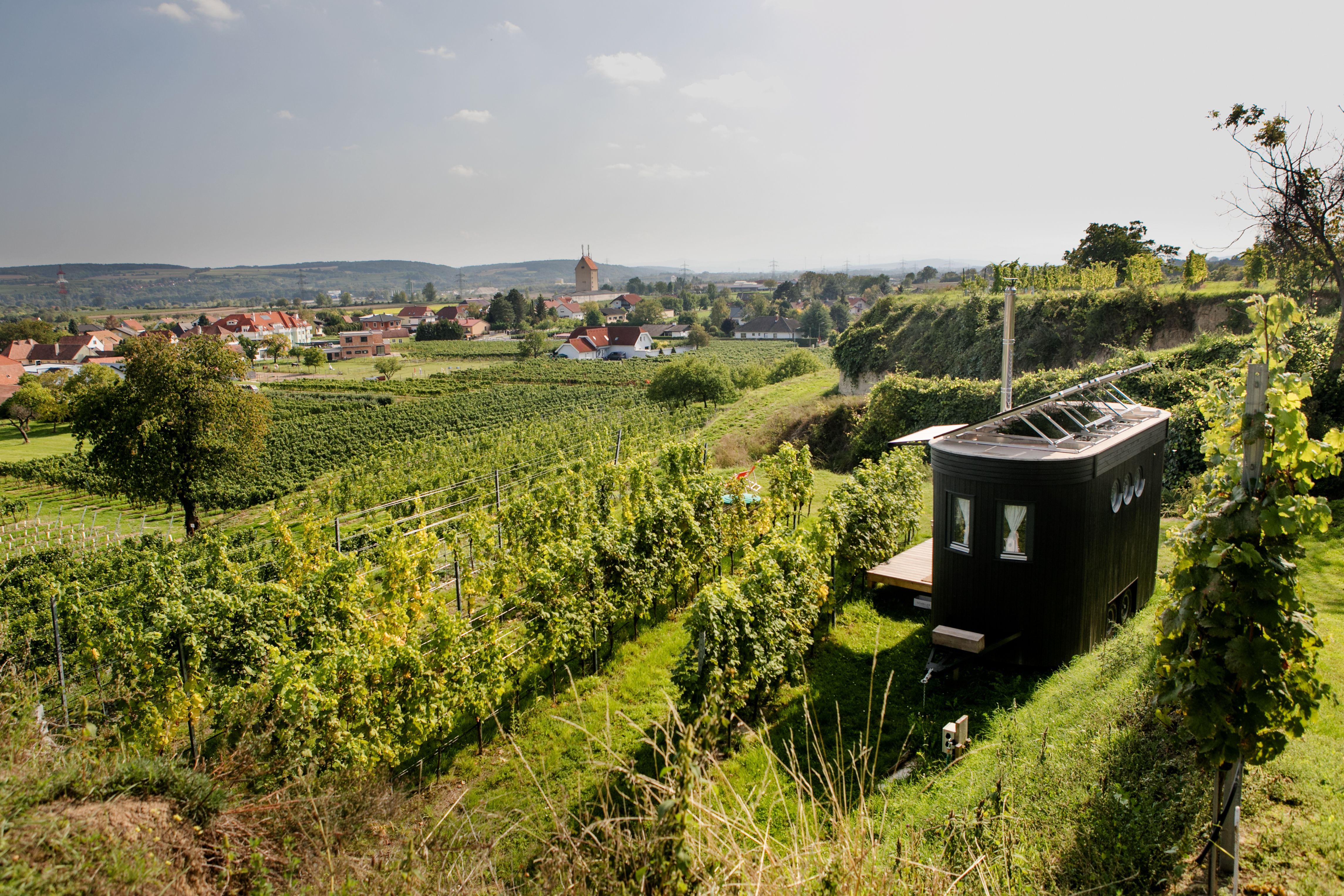A small, modern caravan stands in the middle of vineyards with a view of a village and hills in the background.