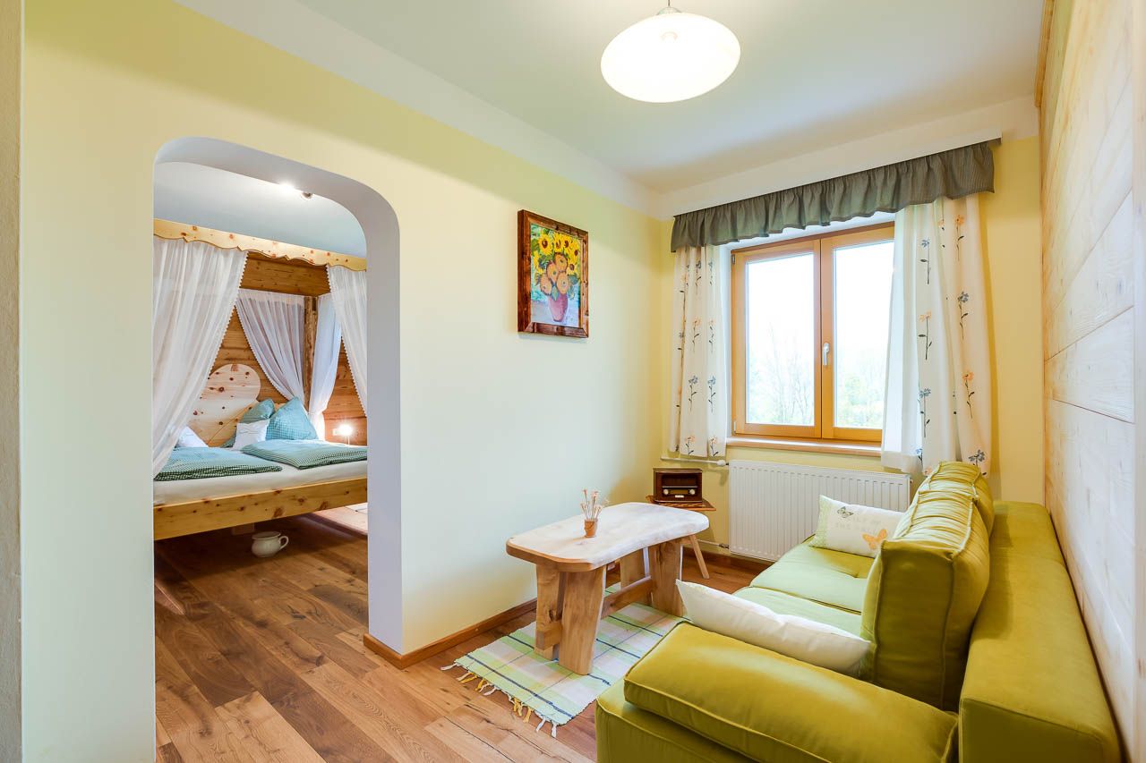 Cozy room with wooden furniture, green sofa, large window and a four-poster bed in the background.
