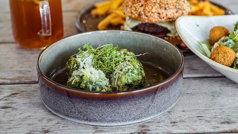 A plate with spinach dumplings, parmesan and cress. In the background, a drink, a burger and a salad. 