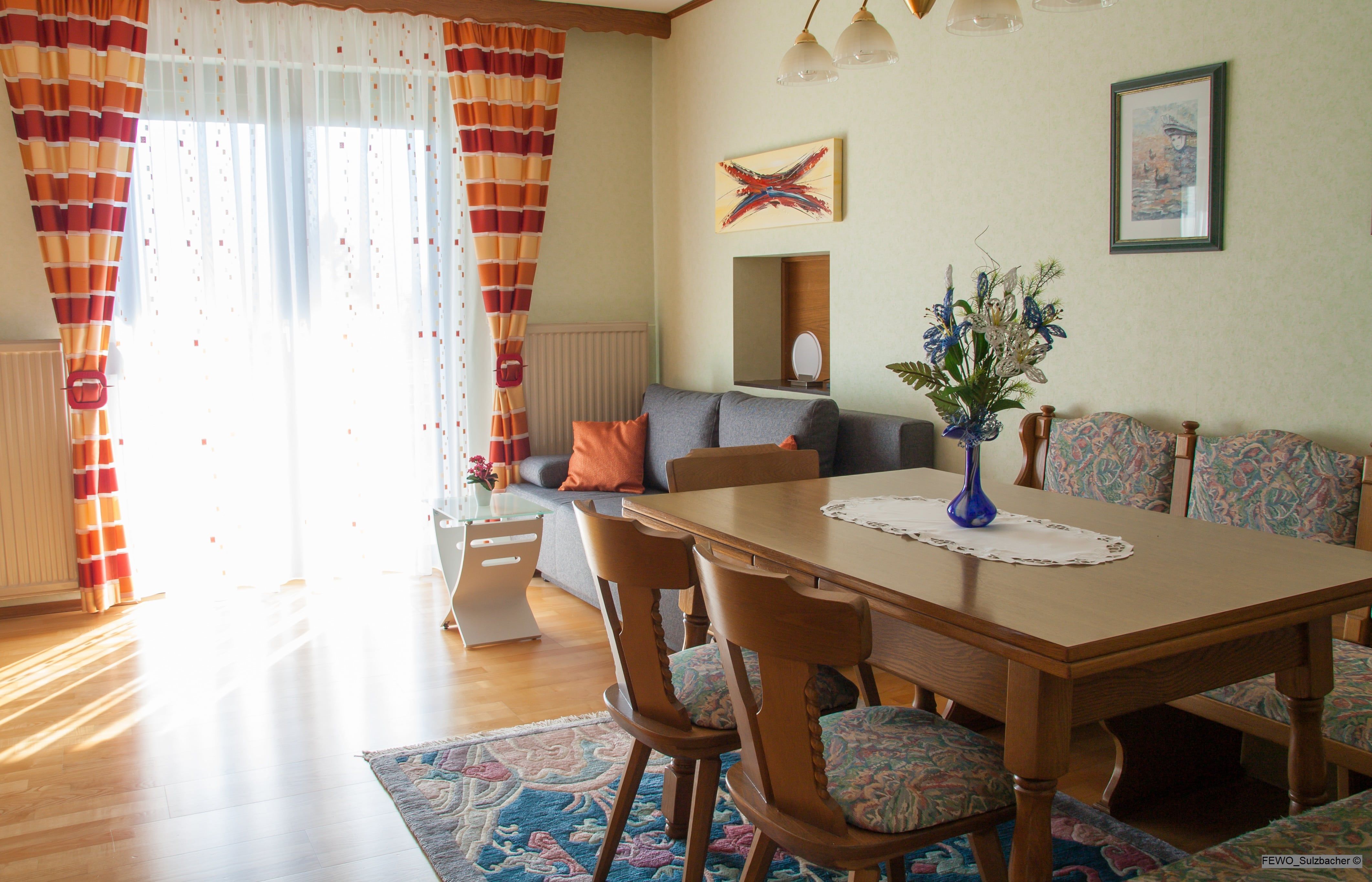 A cozy living room with a wooden table, chairs, a grey sofa and colorful curtains. There is a bouquet of flowers on the table.