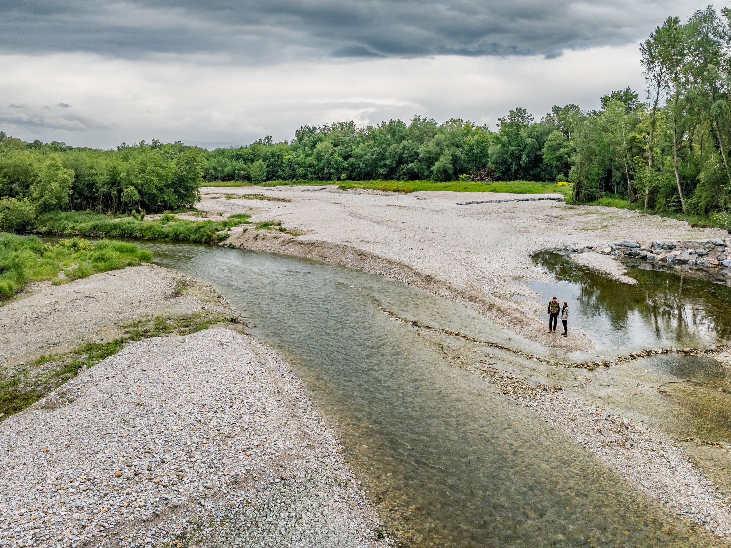 A wide gravel bed of a river with clear water flowing through it. Two people standing by the river, green forest in the background