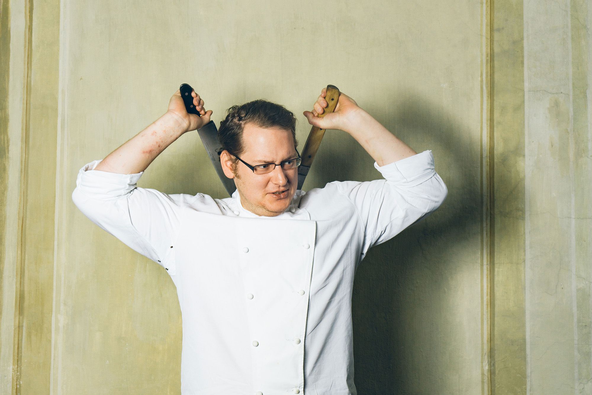A man in chef's clothes holds two knives behind his head.