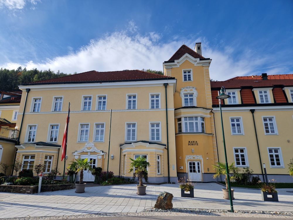 Yellow building with red roofs and several windows, surrounded by plants and flags.