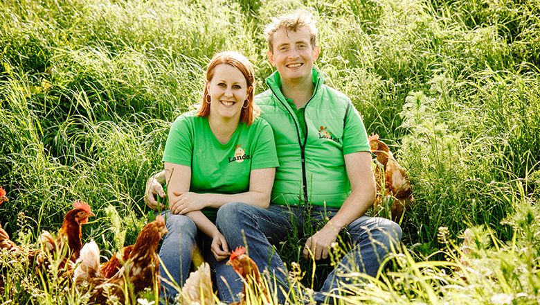 A man and a woman in green T-shirts sit smiling in a meadow surrounded by chickens.