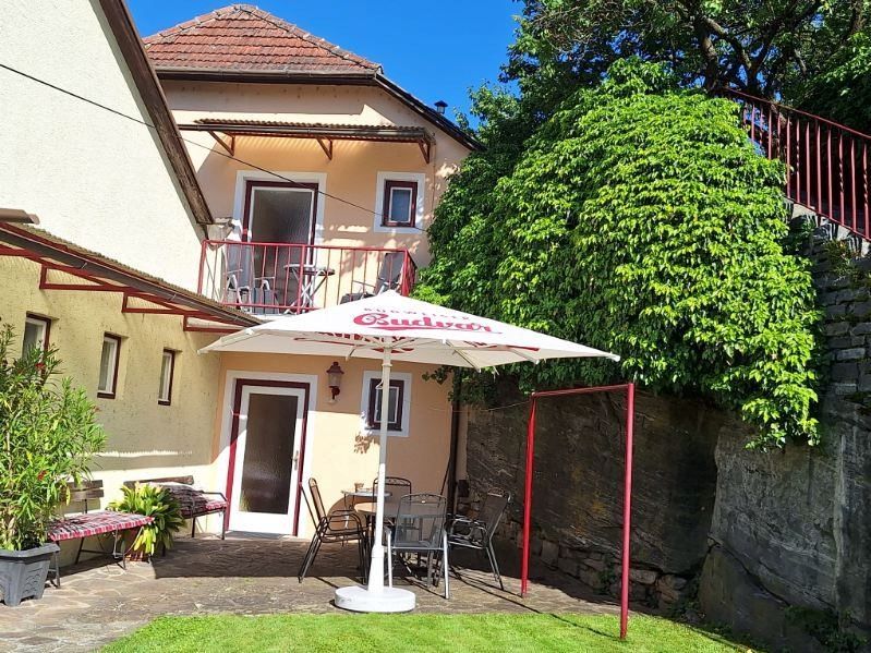 A cozy garden with a parasol, table and chairs in front of a house with a balcony and green climbing plants.