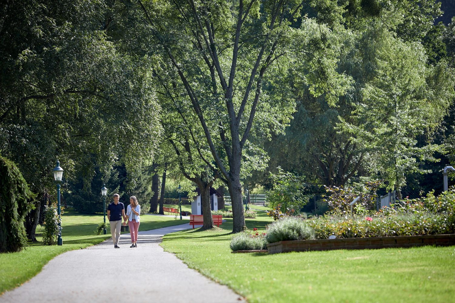 A couple walks along a path in the spa gardens of Bad Schönau, surrounded by green trees and well-tended flowerbeds.