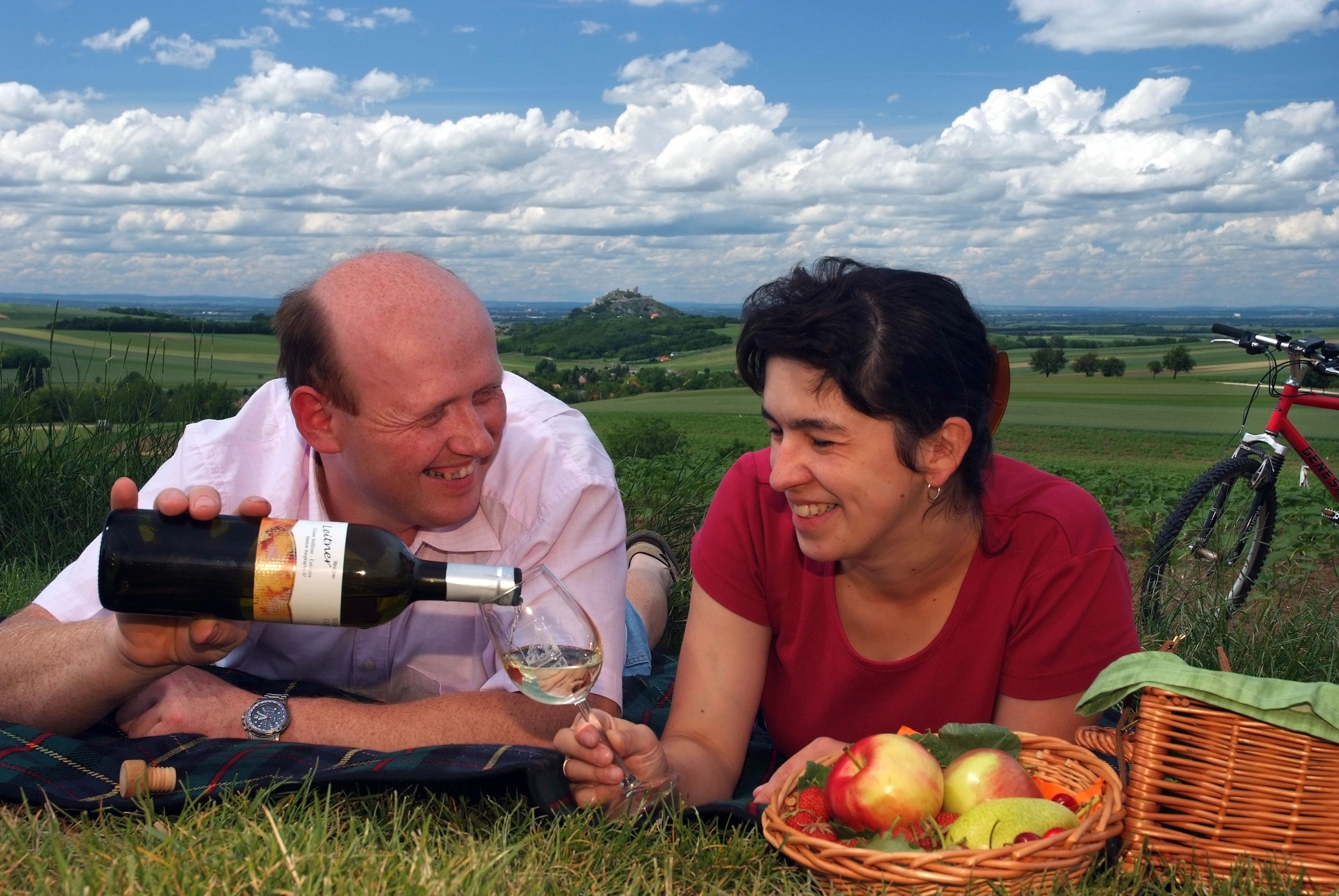 A couple having a picnic in a meadow with wine and a fruit basket, in the background a hilly landscape and a bicycle.