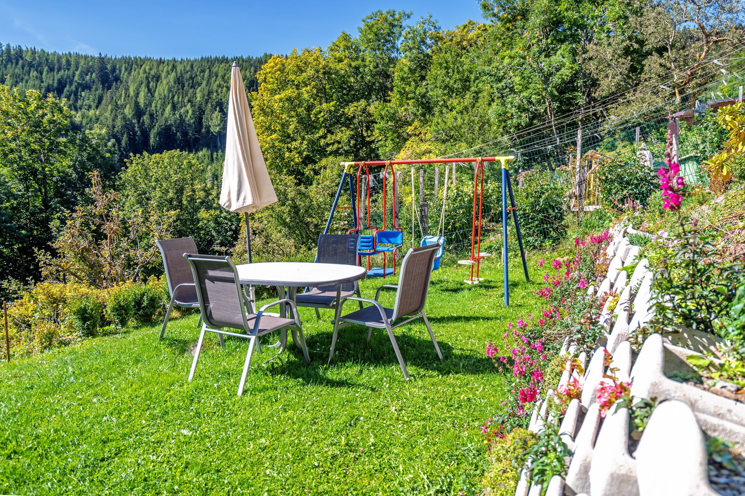 A playground with table, chairs, parasol and swings on a lawn surrounded by trees and flowers.