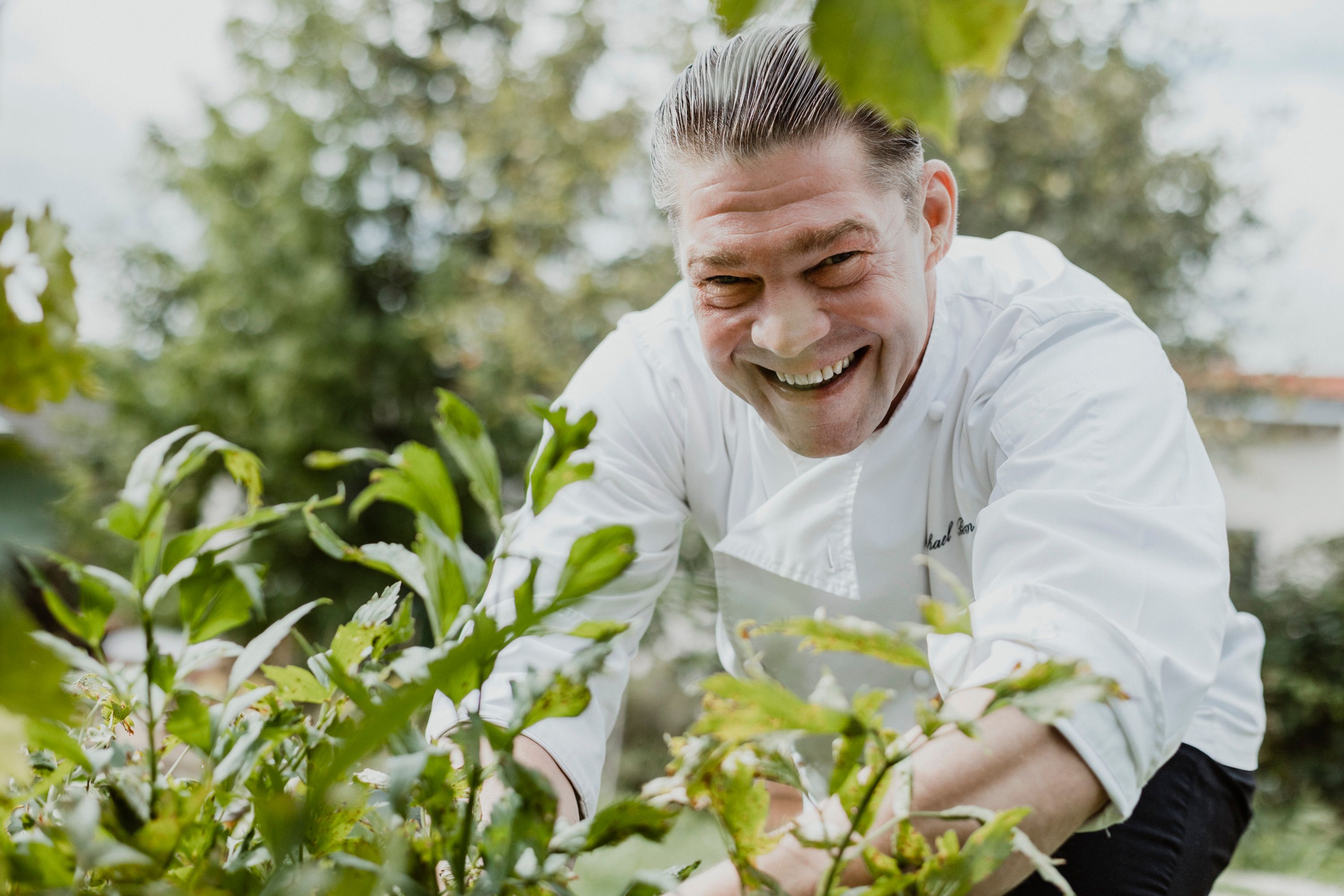 Smiling chef in white uniform in the garden, surrounded by plants.