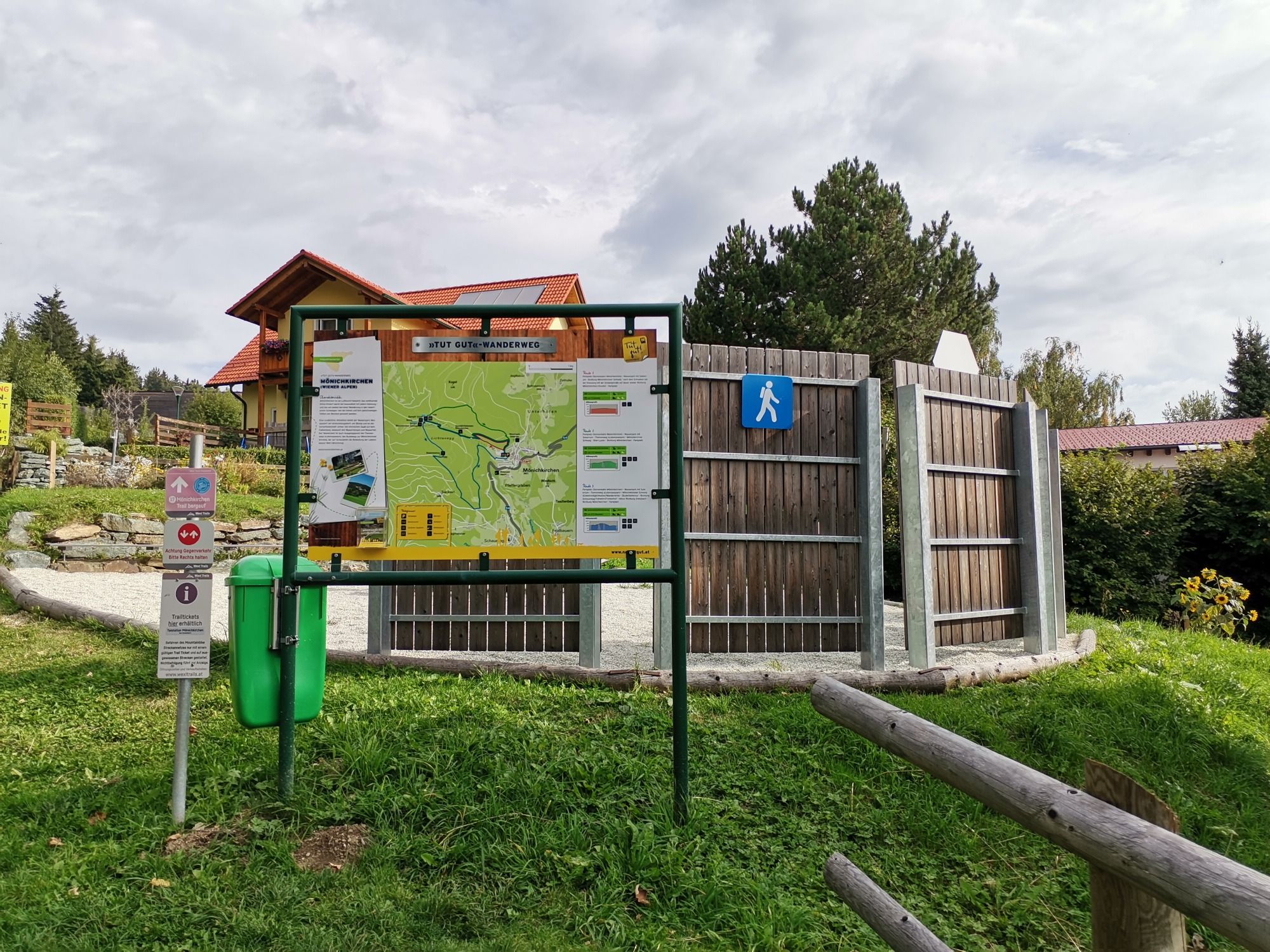 Hiking trail information board with map and signs on a meadow, surrounded by trees and a building in the background.