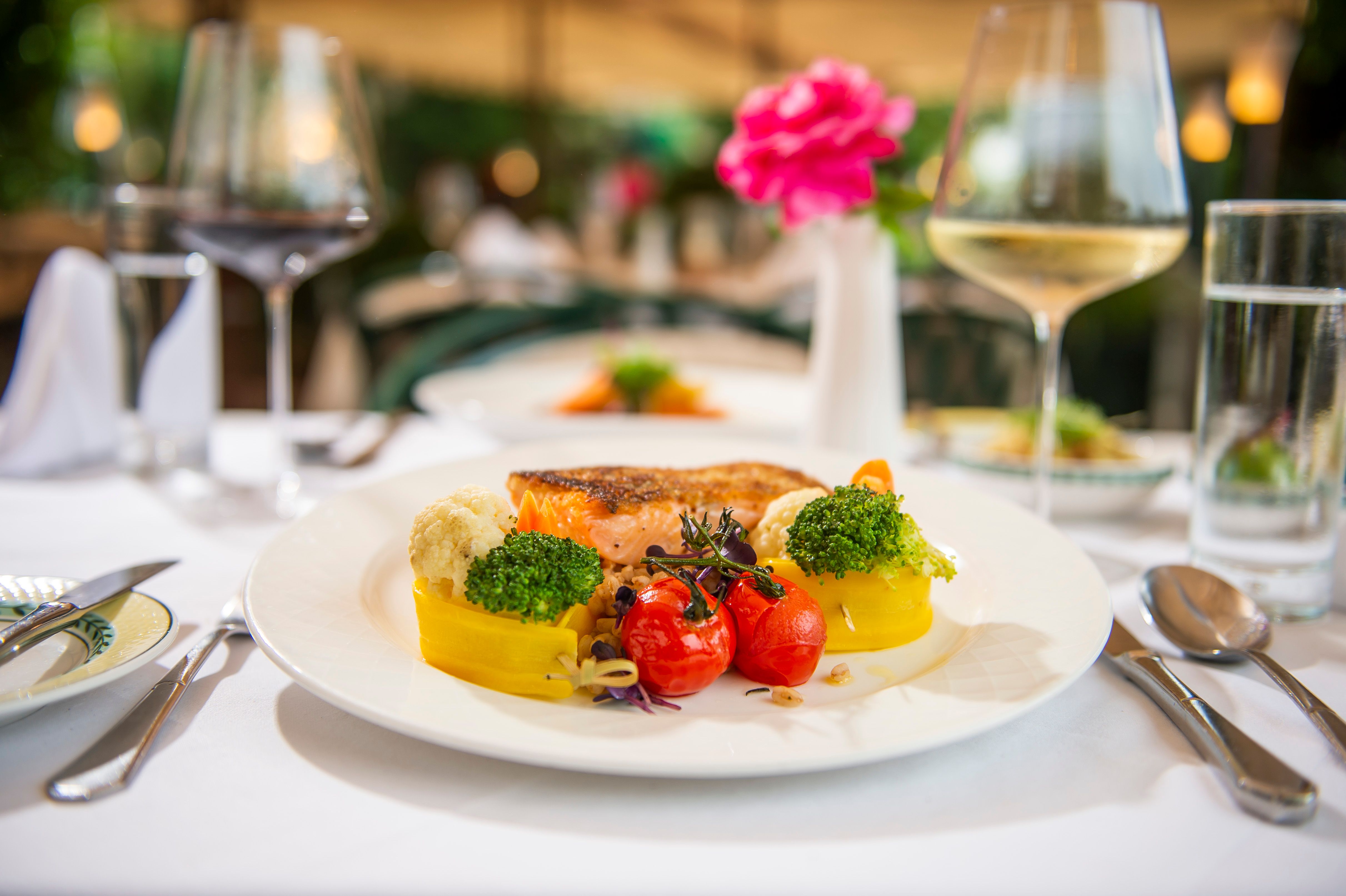 A plate of grilled salmon, broccoli, cauliflower, yellow peppers and cherry tomatoes, on a table set with wine glasses.