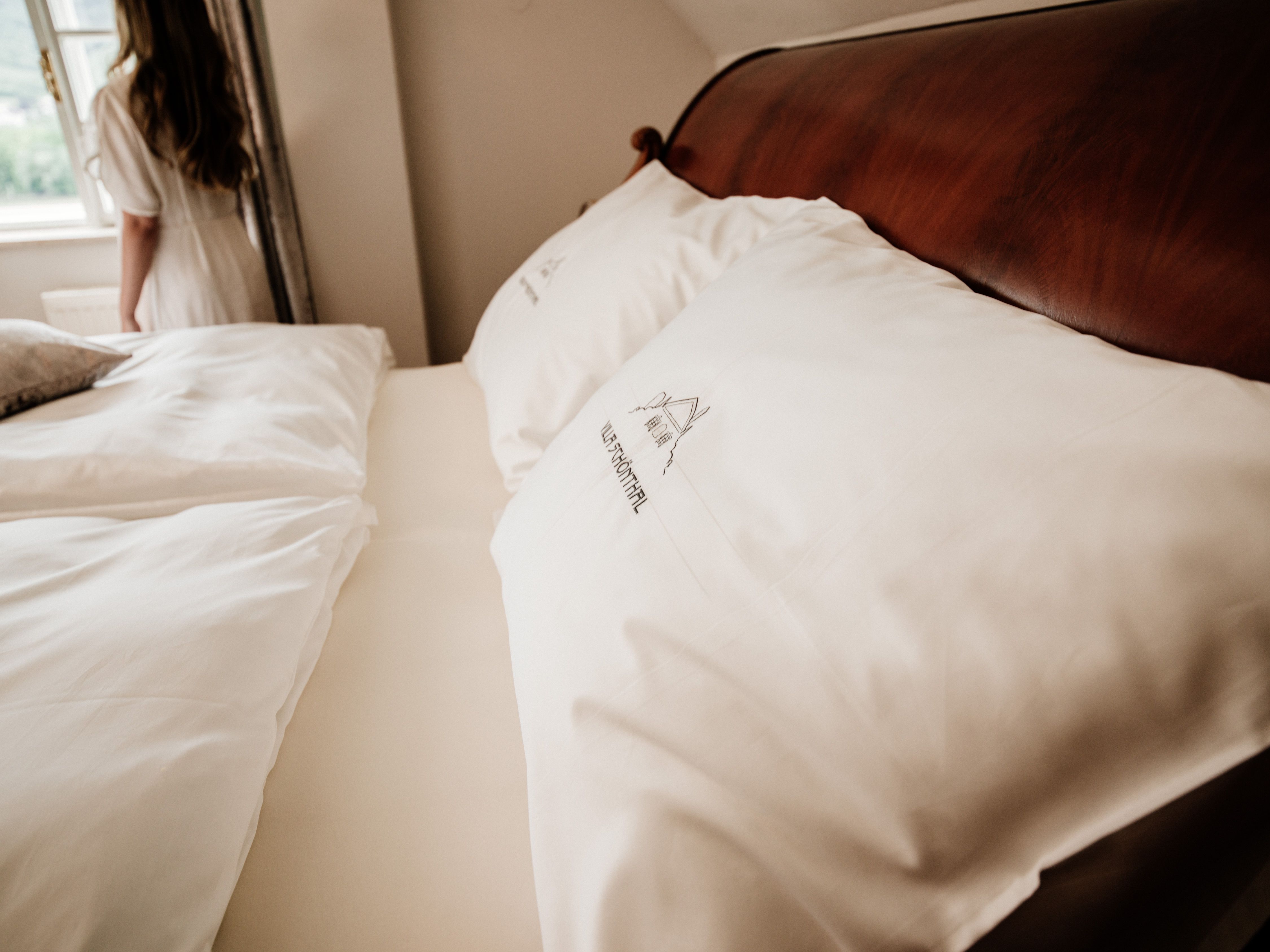 Close-up of a bed with white pillows and bed linen, in the background a woman at the window.
