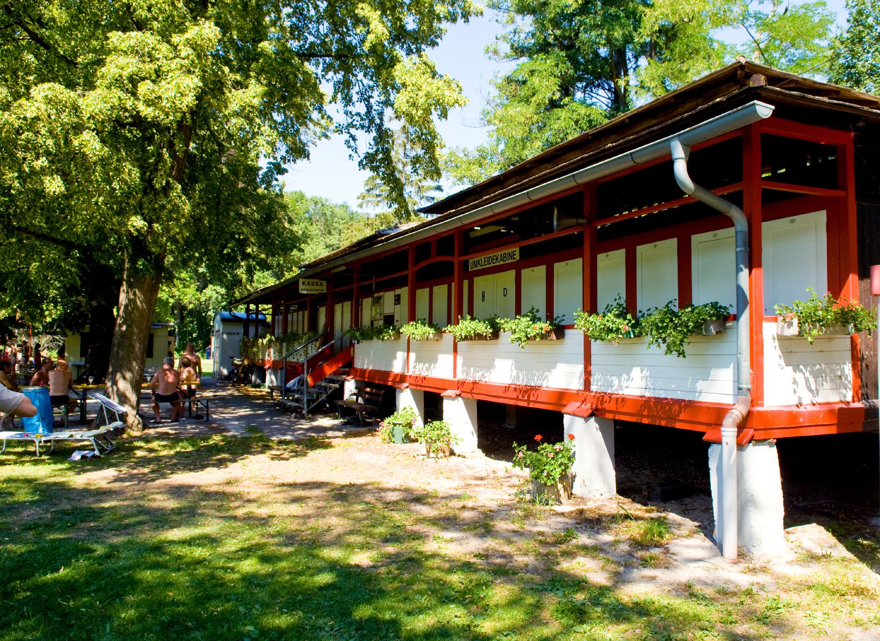 A red building with changing rooms and plants in front of it, surrounded by trees and people on benches.