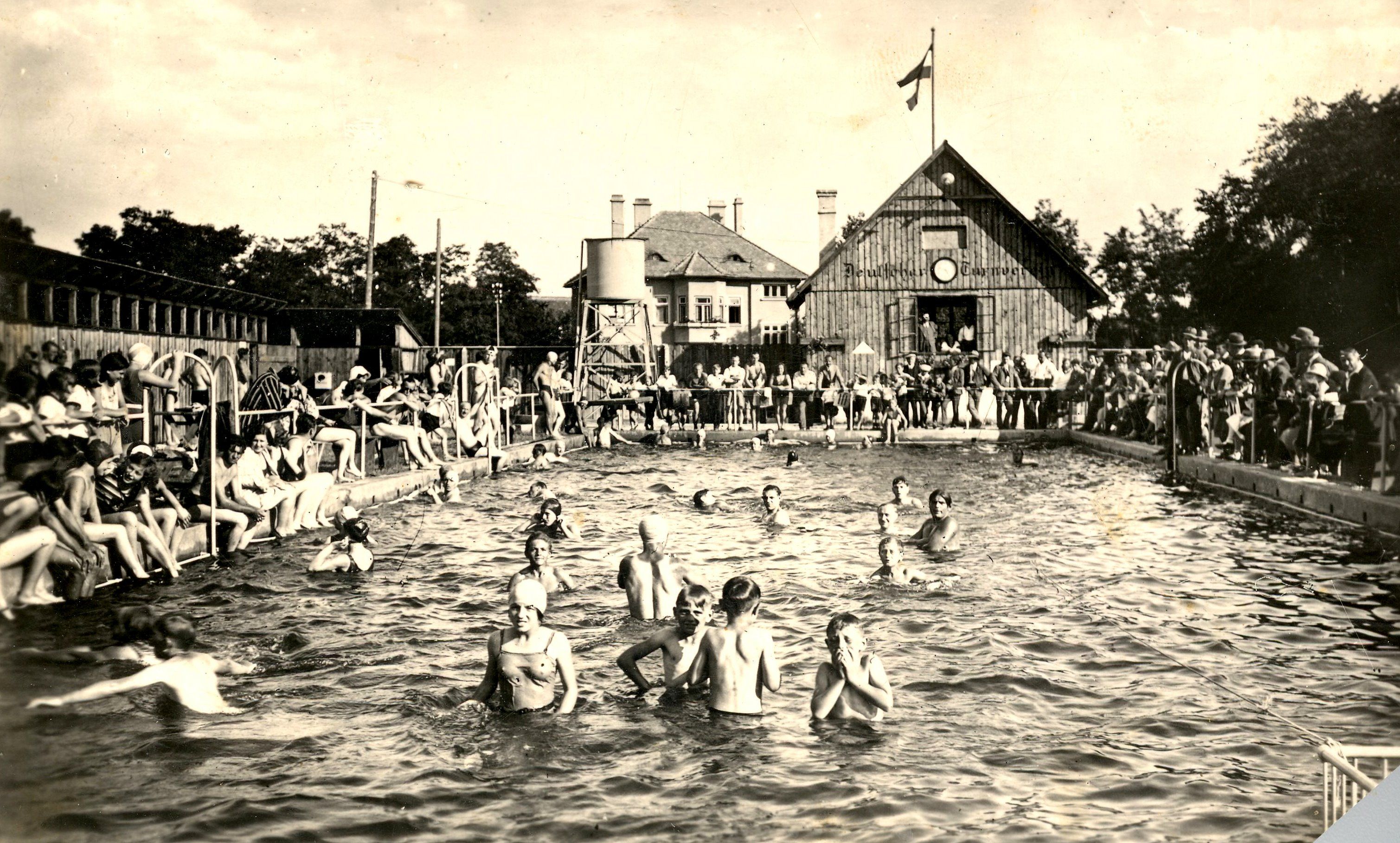 A historical photo of an outdoor swimming pool with many people in the water and at the edge of the pool. In the background is a building with a flag.