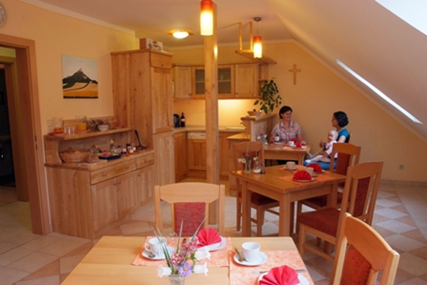 A cozy breakfast room with wooden furniture, laid tables and two people chatting. A child is sitting on one person's lap.