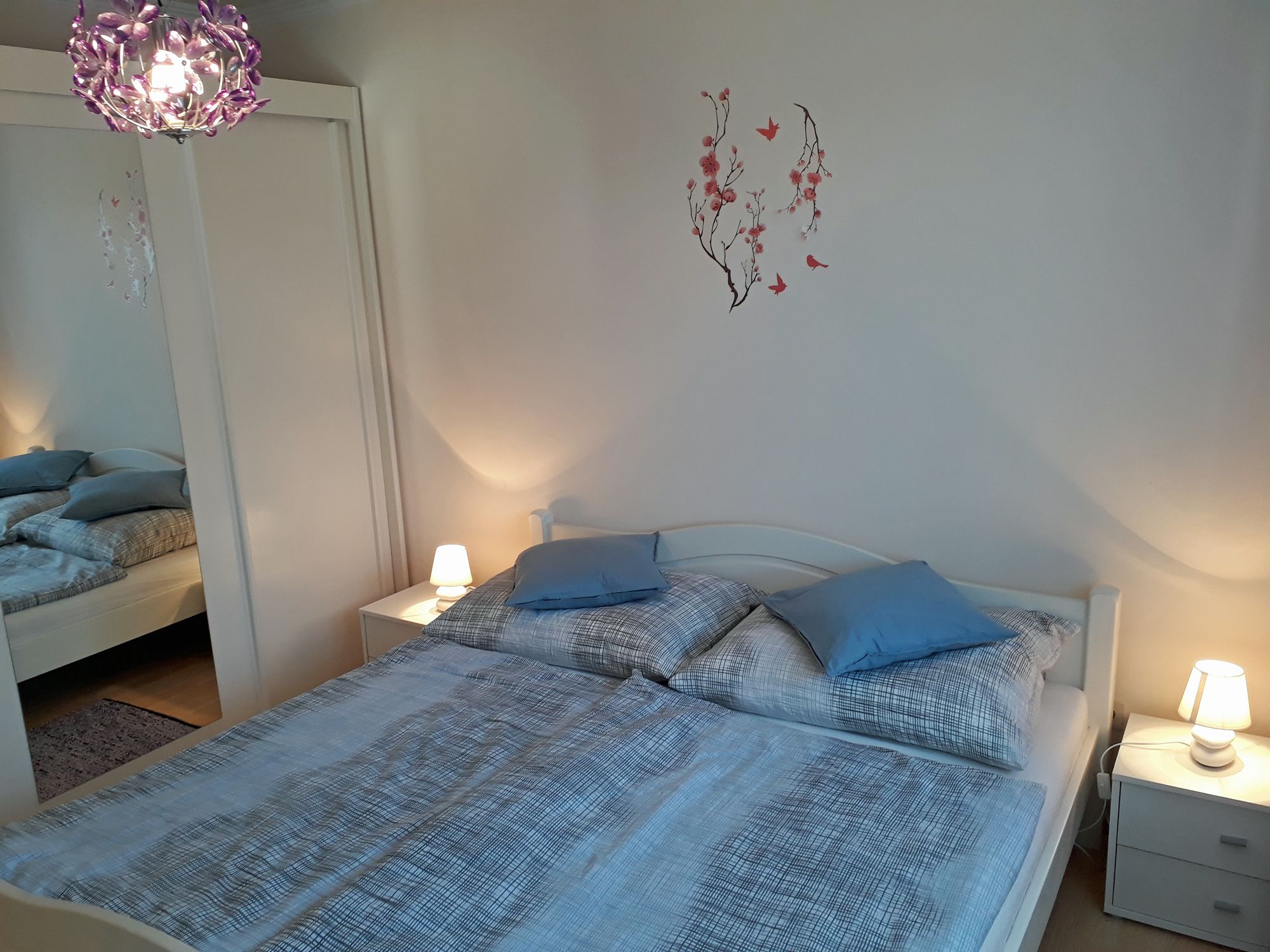 Cozy bedroom with double bed, blue bed linen, bedside tables with lamps and a wall tattoo with flowers and birds.