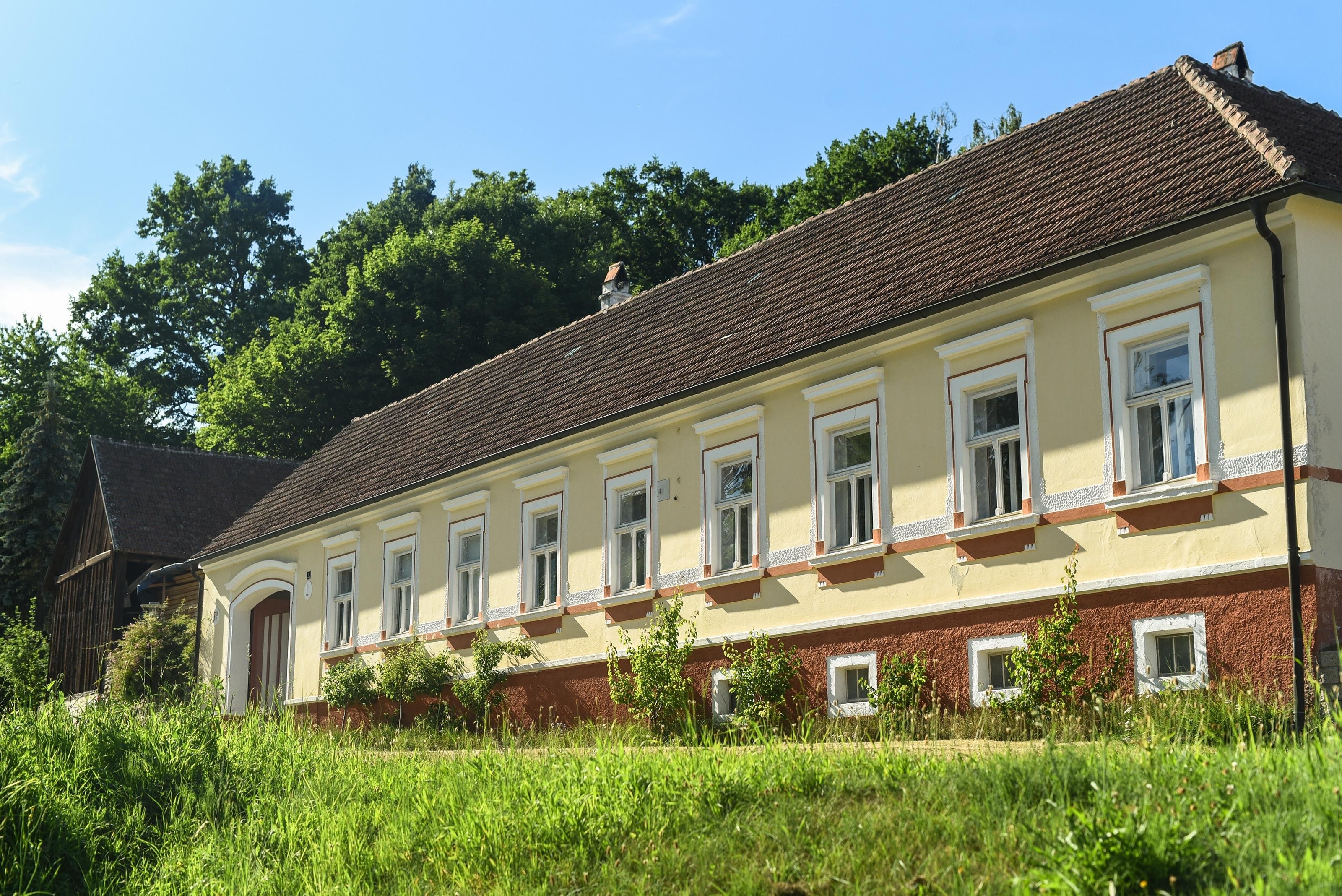 A long, yellow country house with red roof tiles and white window frames, surrounded by greenery and trees in the background.