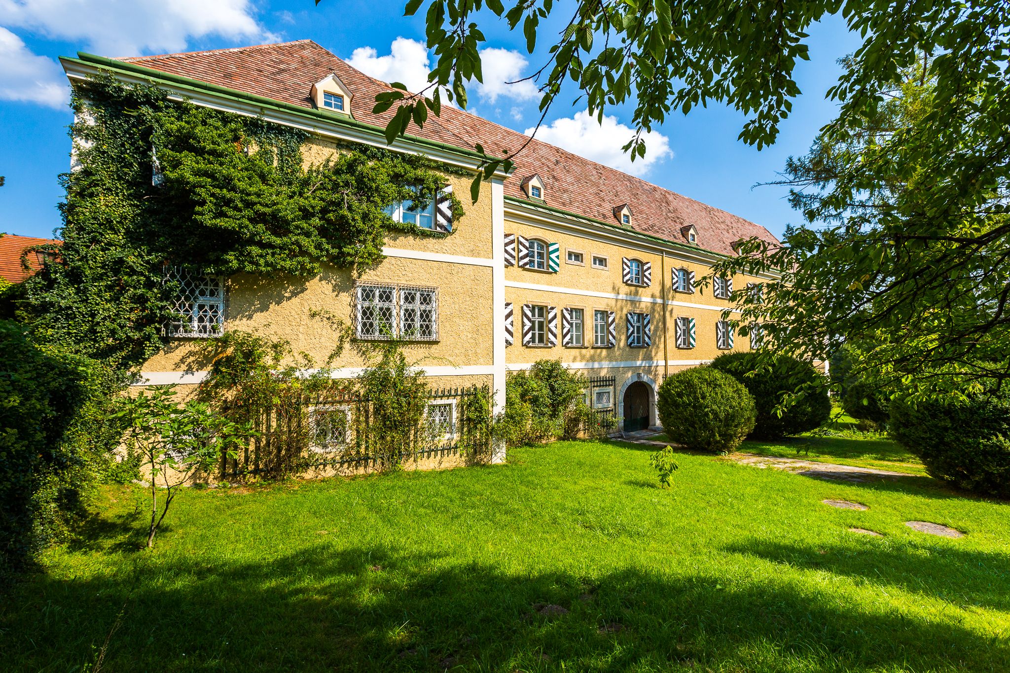 Historic building with yellow façade and ivy growth, surrounded by green meadow and trees.