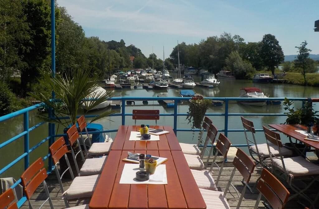 Terrace with wooden tables and chairs, view of a river with boats.