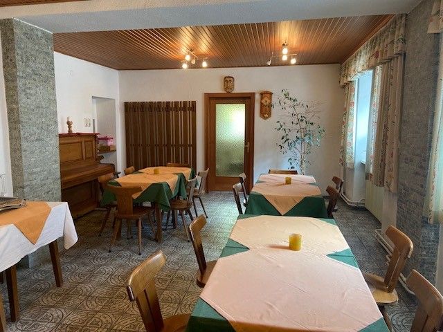 A cozy breakfast room with wooden tables, chairs and a piano. The tables are covered with tablecloths and there are windows with curtains.