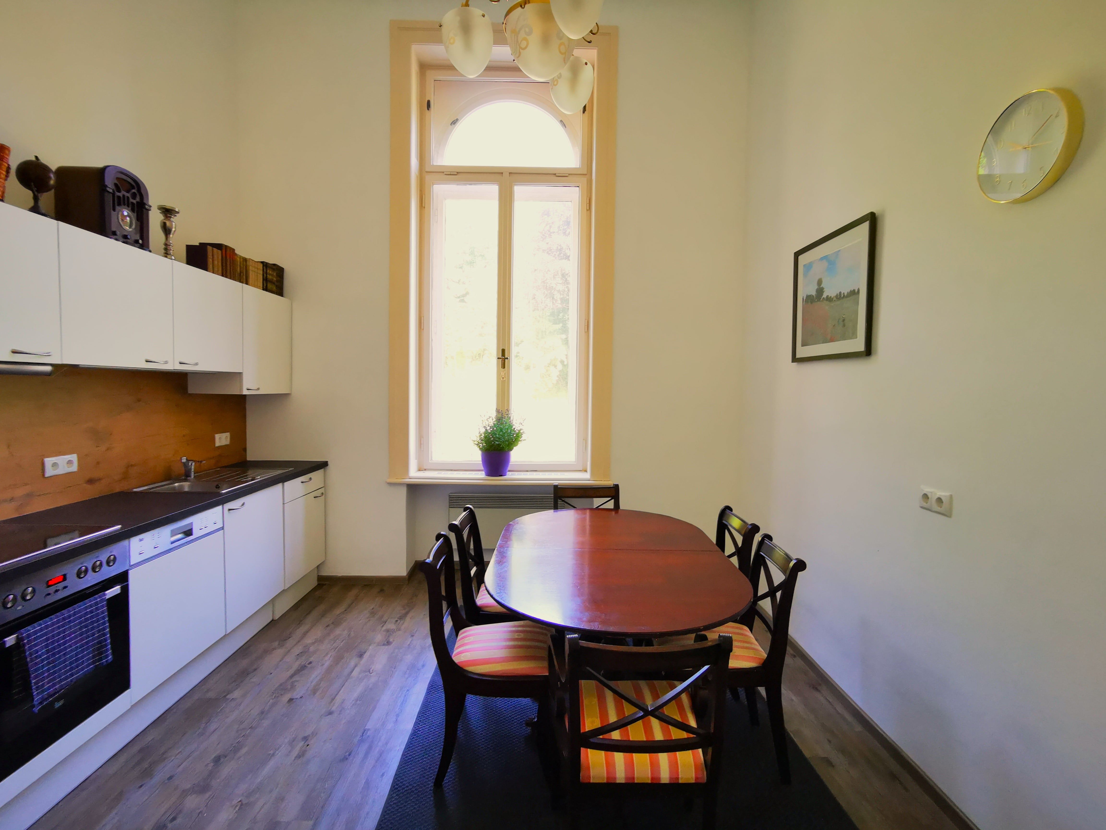 Modern kitchen with dining table and chairs, large window, white cupboards and wooden floor.