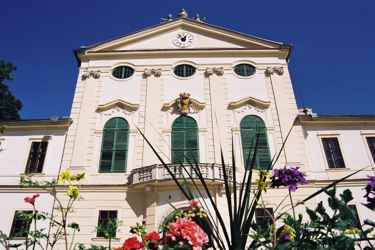 Historic building with green shutters and a clock on the façade, surrounded by colorful flowers.