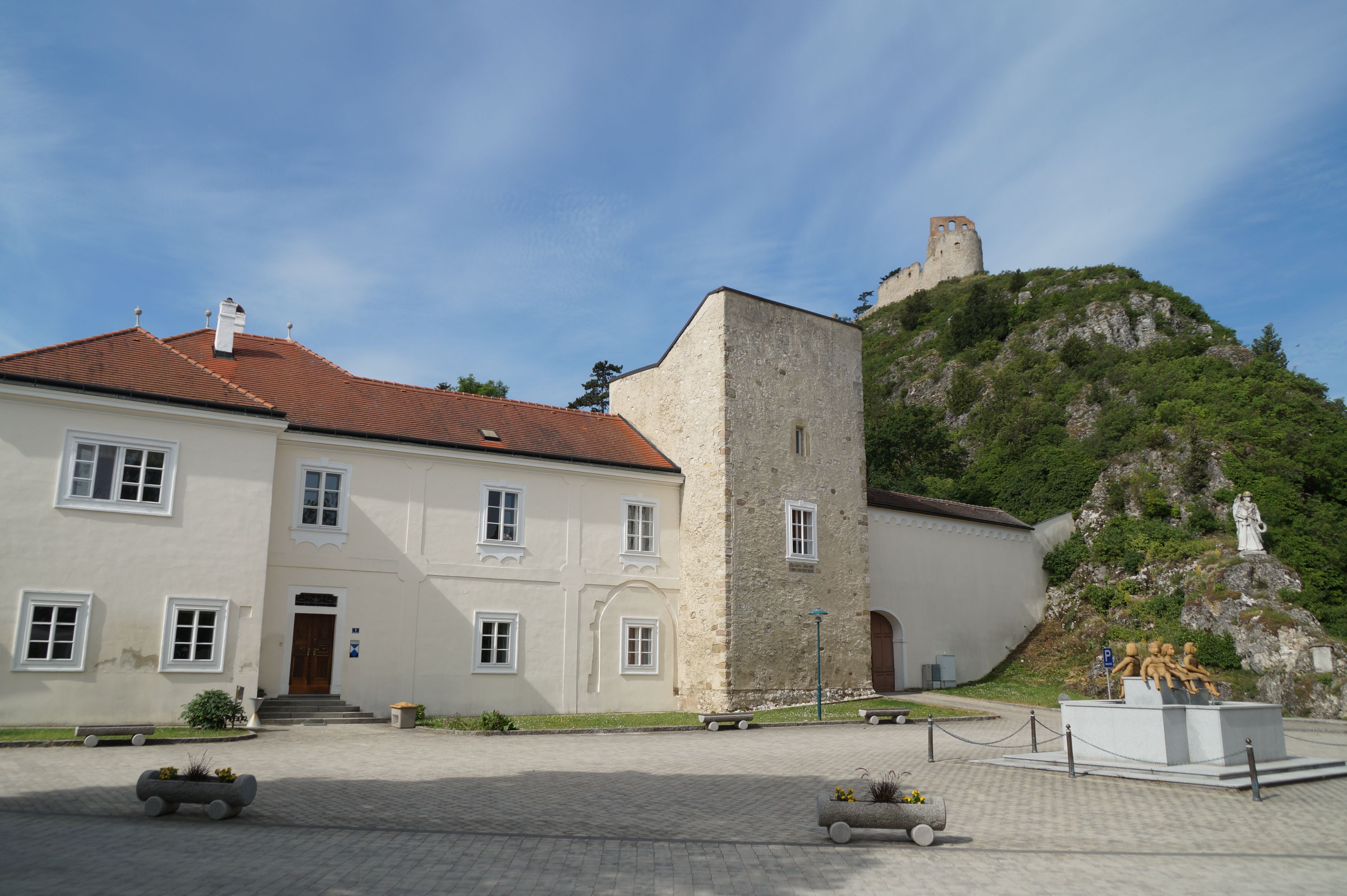 A historic building with a red tiled roof and a stone tower, with a ruined castle on a hill in the background.
