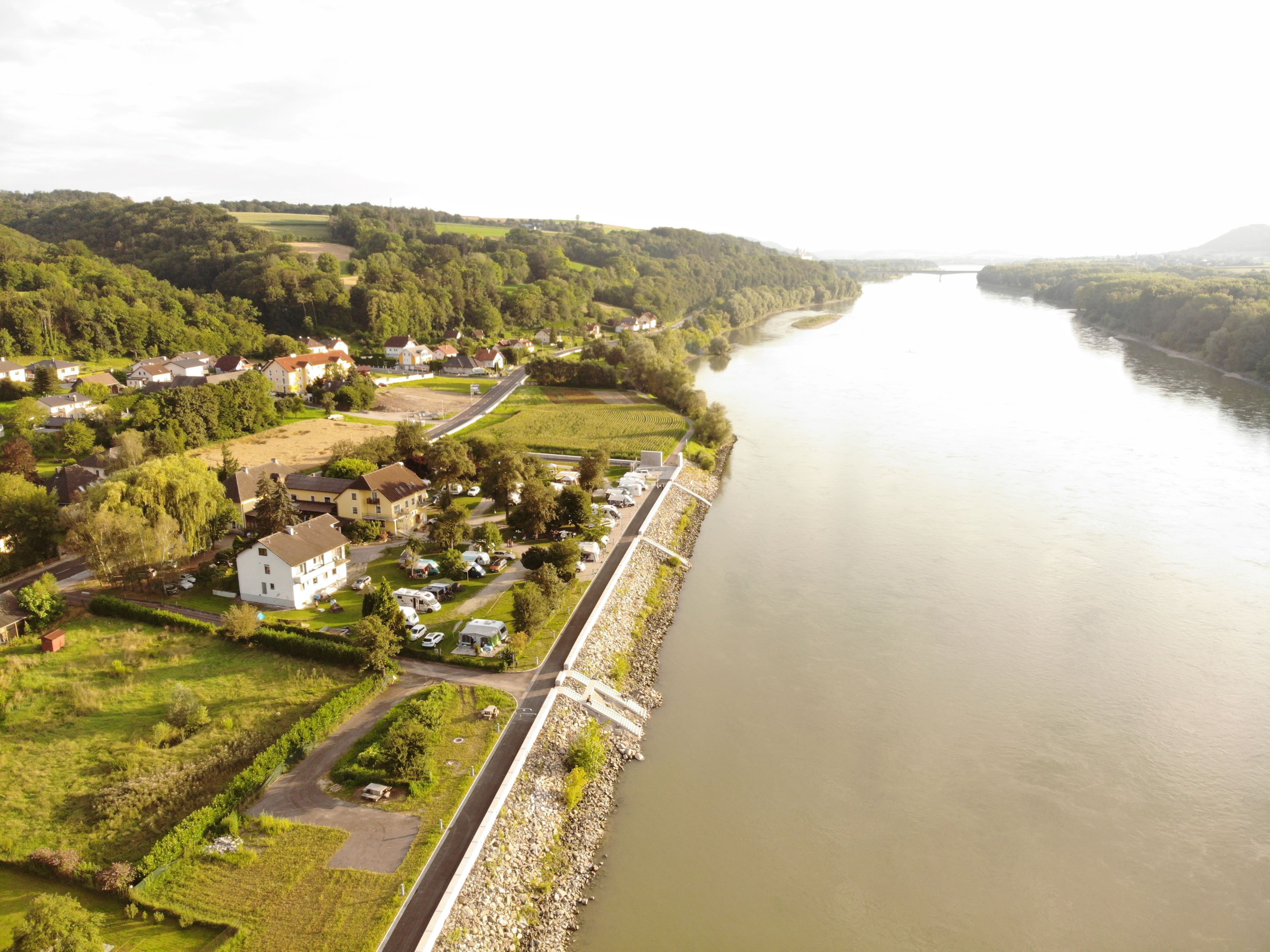 Aerial view of a riverbank with houses and mobile homes, surrounded by green countryside.