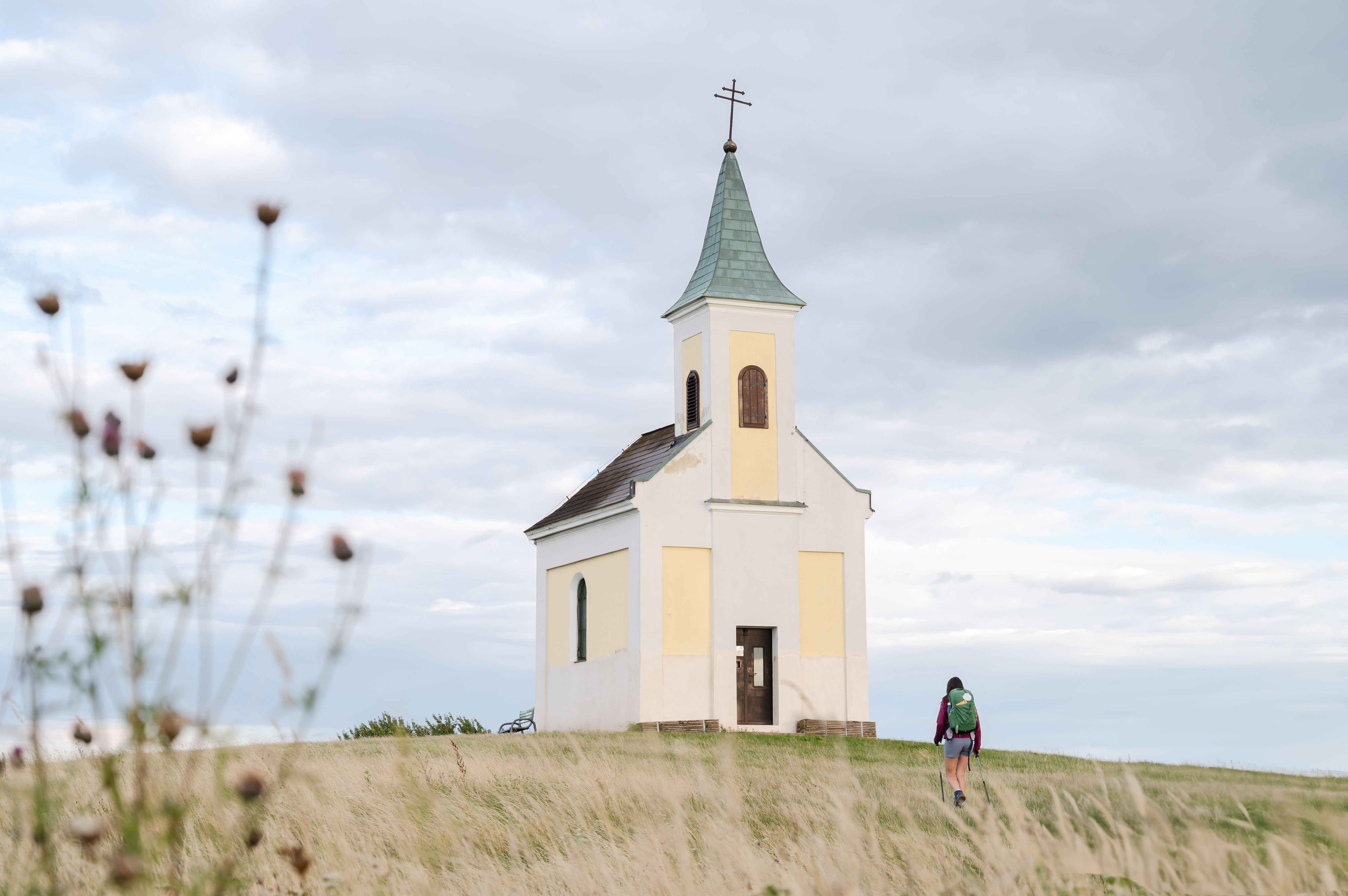 A small chapel on a hill with a person walking towards it, surrounded by a meadow and a cloudy sky.