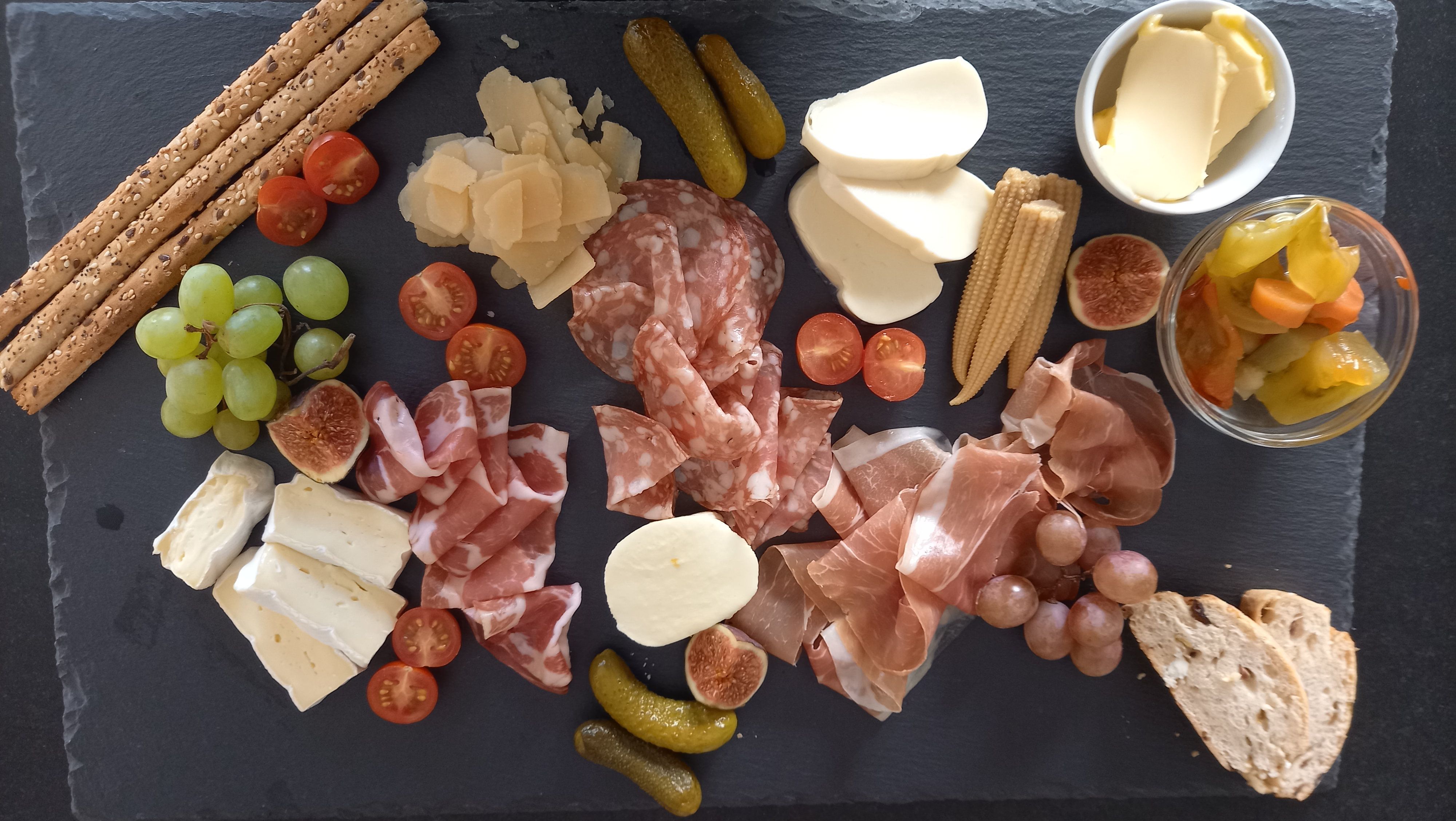 A selection of finger food on a slate platter, including cheese, sausage, grapes, tomatoes, cucumber and bread.