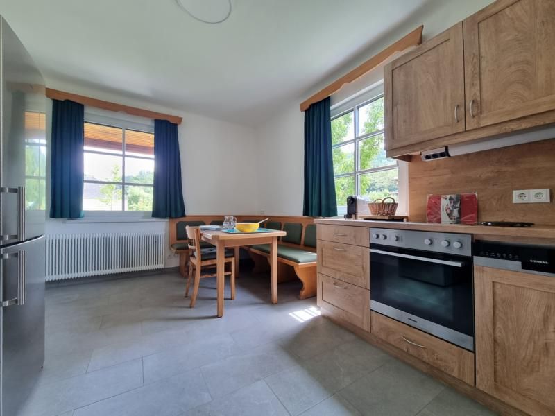 Kitchen with wooden furniture, dining table and corner bench, two windows with green curtains.